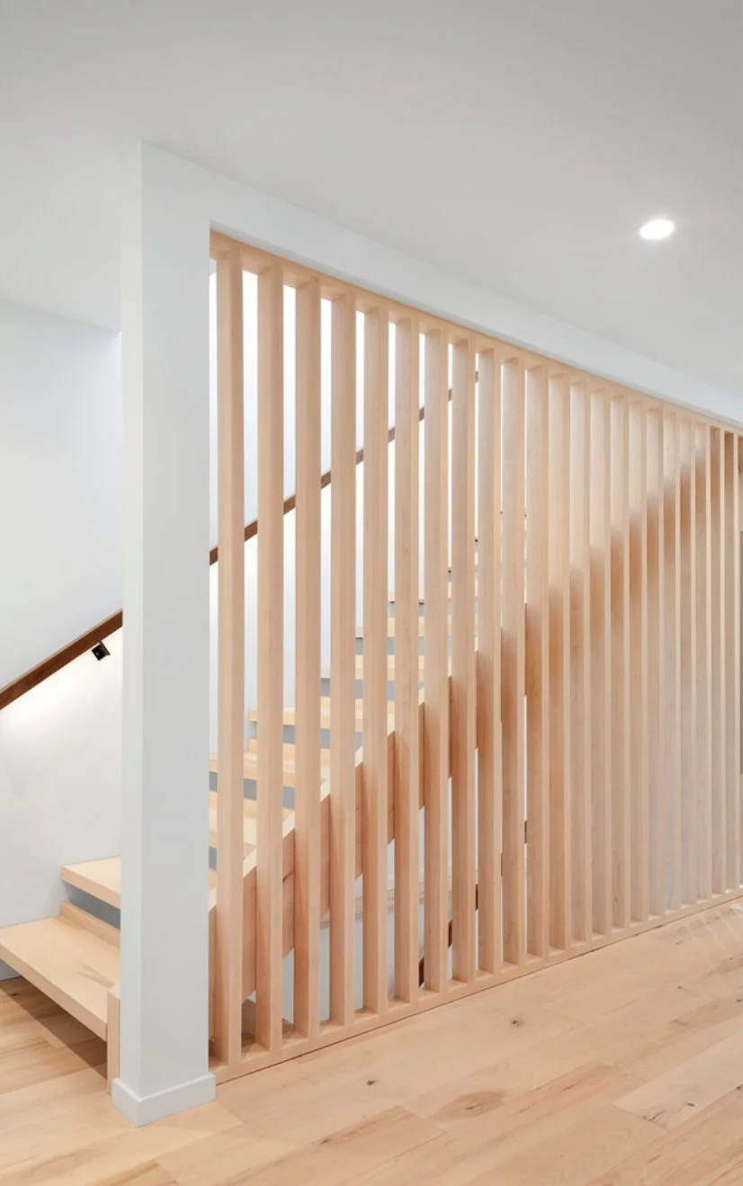 Interior view of a custom space divider architectural feature on a staircase with vertical wooden slats forming a railing, light wood flooring, and white walls.