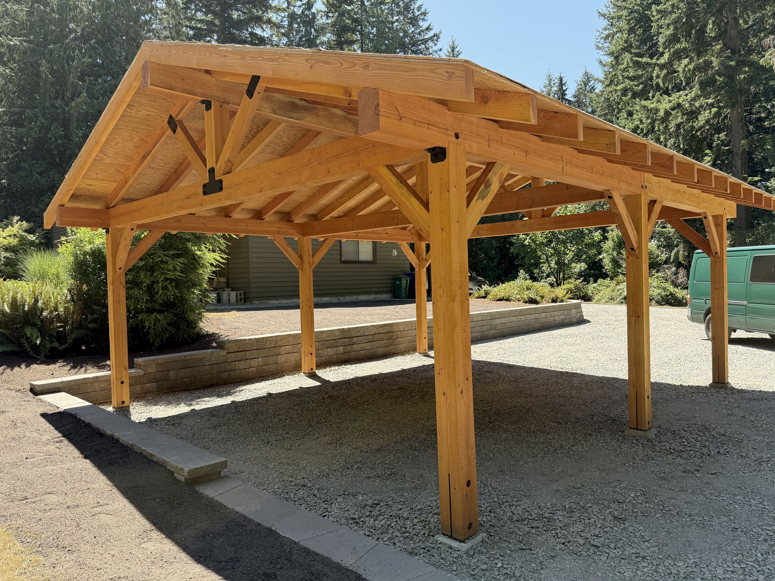 Wooden carport structure with a pitched roof situated on gravel and surrounding a landscaped area with plants and trees.
