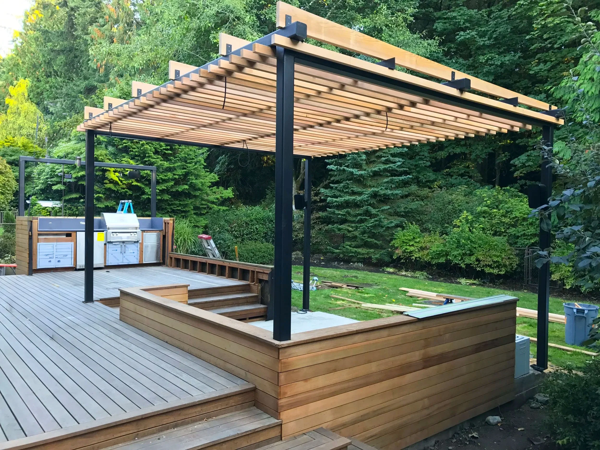 A newly constructed custom multi level outdoor wooden deck with cooking area, pergola, and lush green trees in the background, undergoing finishing touches.