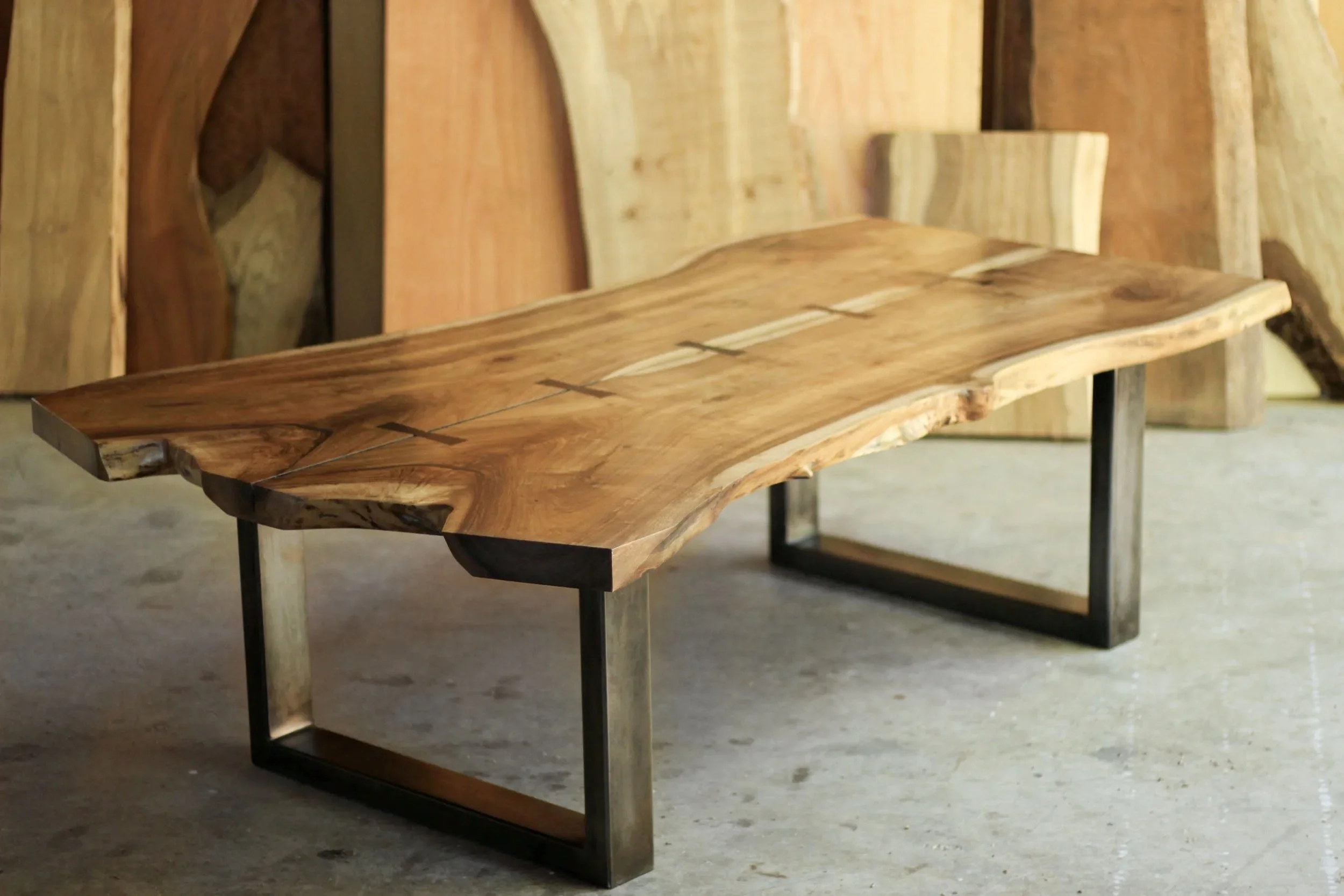 A wooden live edge table with metal legs in a woodworking shop, with wood slabs and boards in the background.