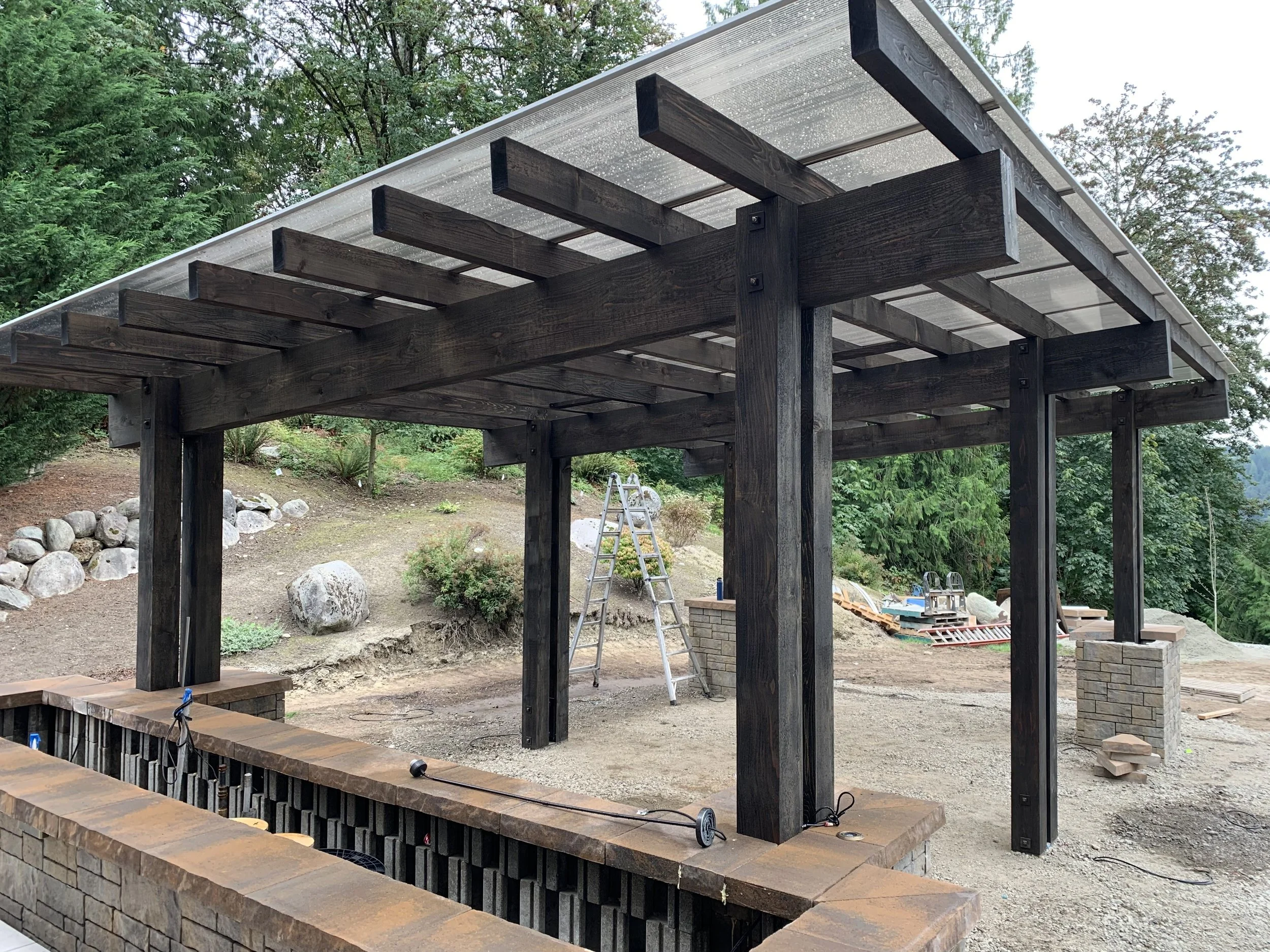 Construction of a wooden pergola with dark stained beams and a clear plastic roof in a backyard, with trees and rocks in the background.