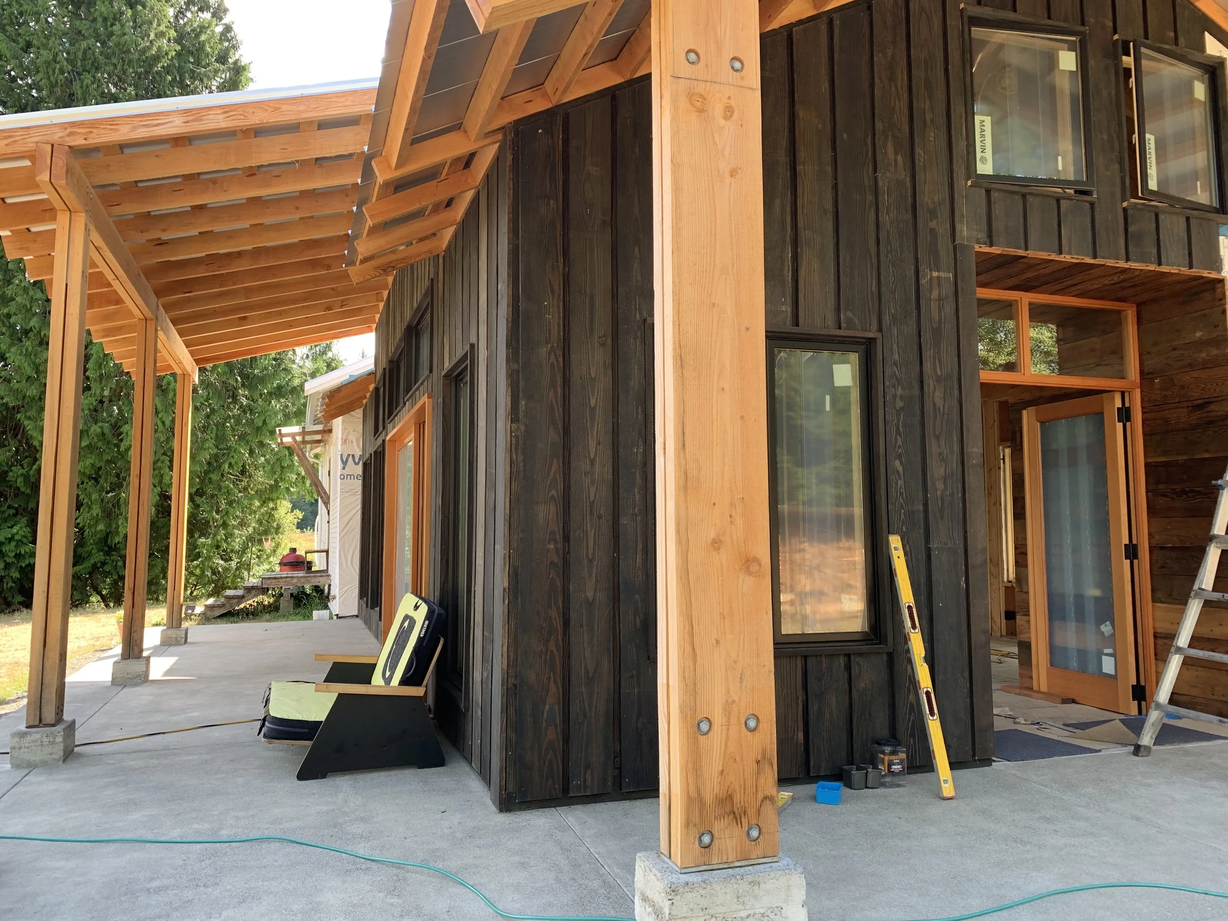 Under construction pergola on modern house with black wood siding, large windows, and a wooden porch with a roof.