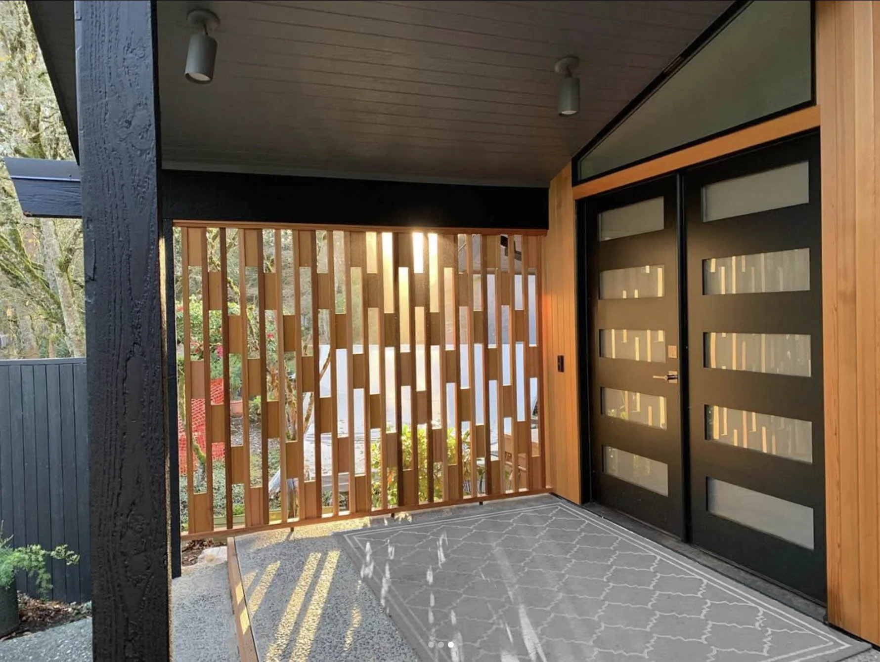 Modern front porch custom privacy screen woodwork with black double doors, and a gray patterned rug, sunlight streaming through trees.
