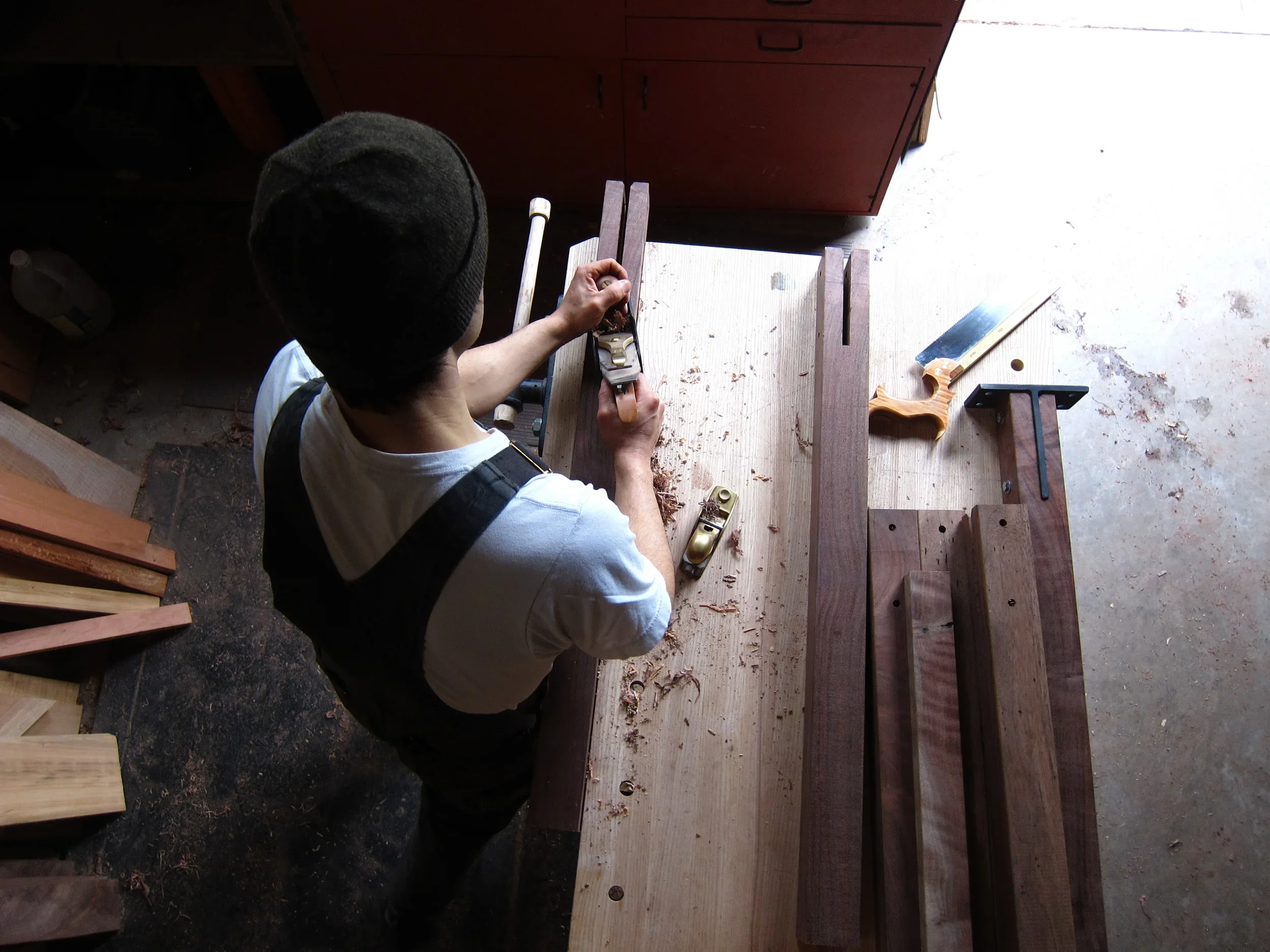 Overhead view of a person working on a woodworking project at a workbench, using a hand plane to smooth a piece of wood. Various woodworking tools and pieces of wood are on the workbench.