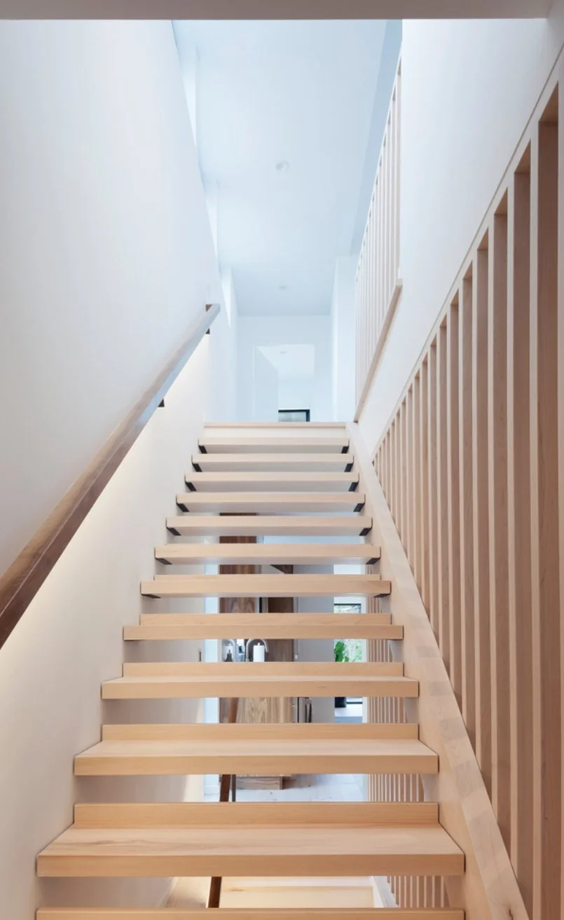 Wooden staircase with open risers in a modern home, viewed from the bottom looking up toward the second floor with natural light flooding the space.