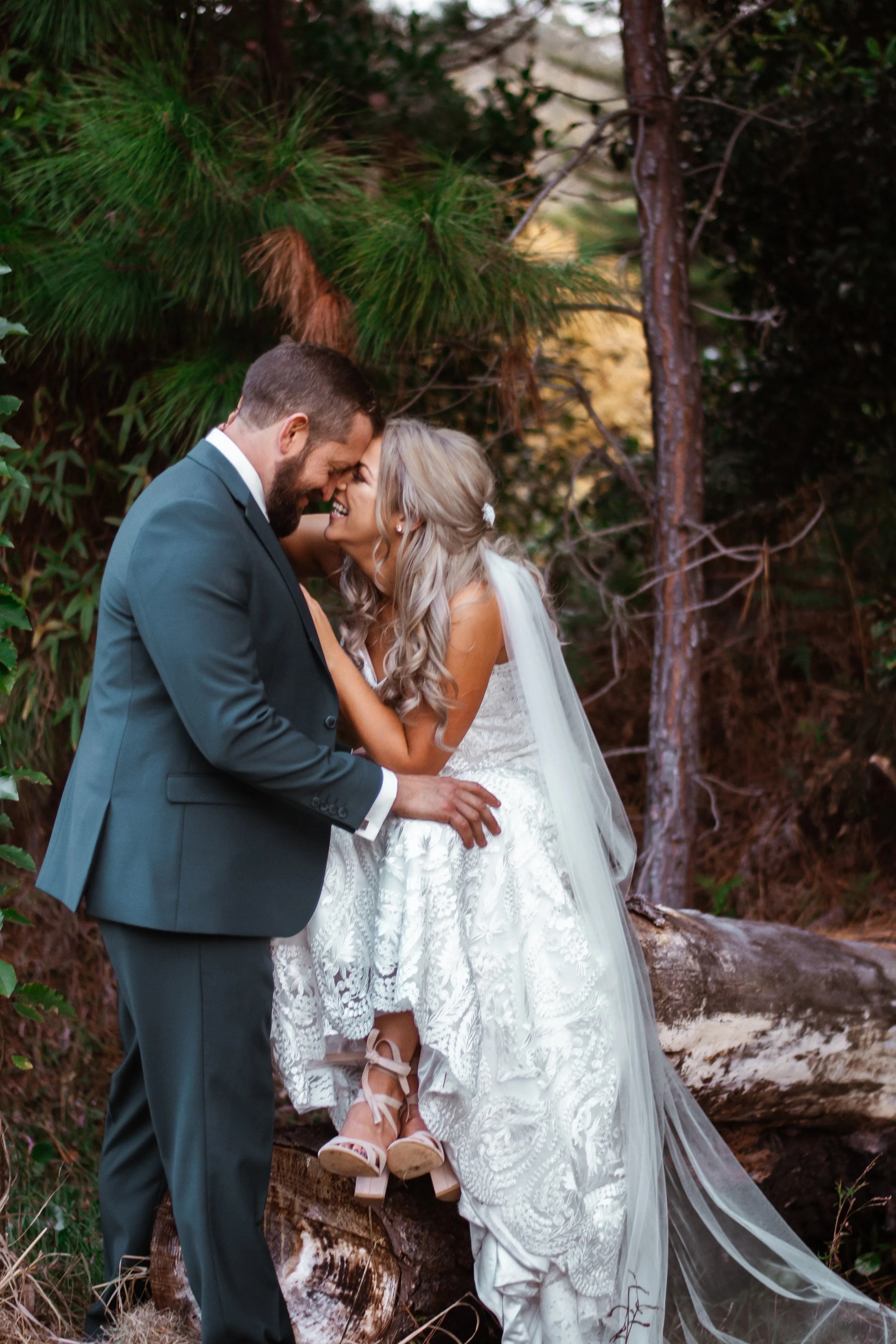 A bride and groom share a joyful moment during their outdoor wedding ceremony, standing on a wooden log with scenic trees in the background.