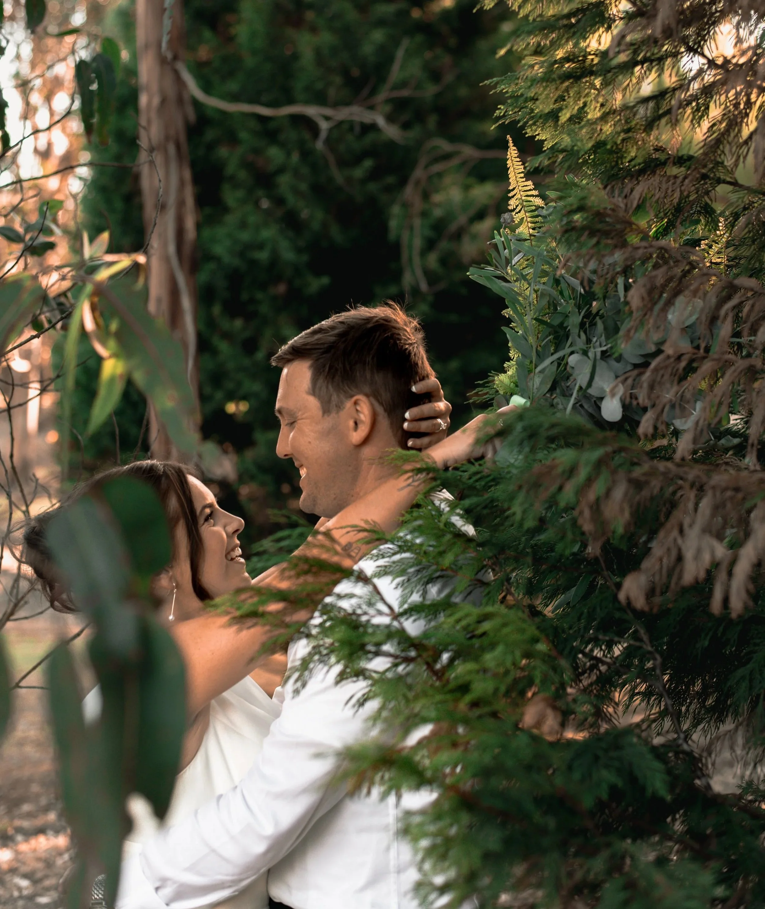 A couple in white clothing sharing a kiss amid lush green foliage, with warm sunlight filtering through the trees in the background.