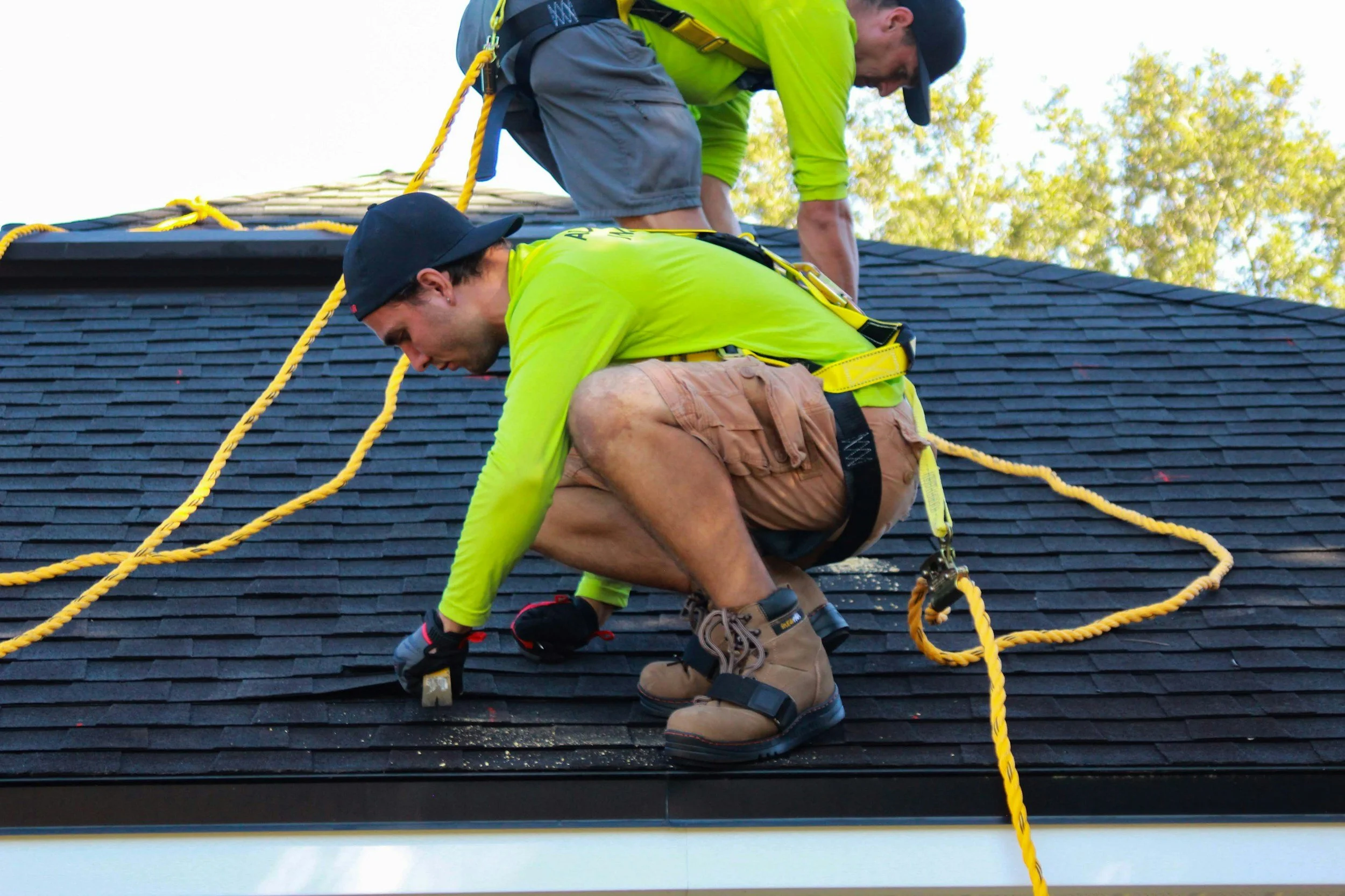 Two workers in bright yellow shirts are installing or repairing a black shingle roof, kneeling and crouching while handling tools and safety ropes.