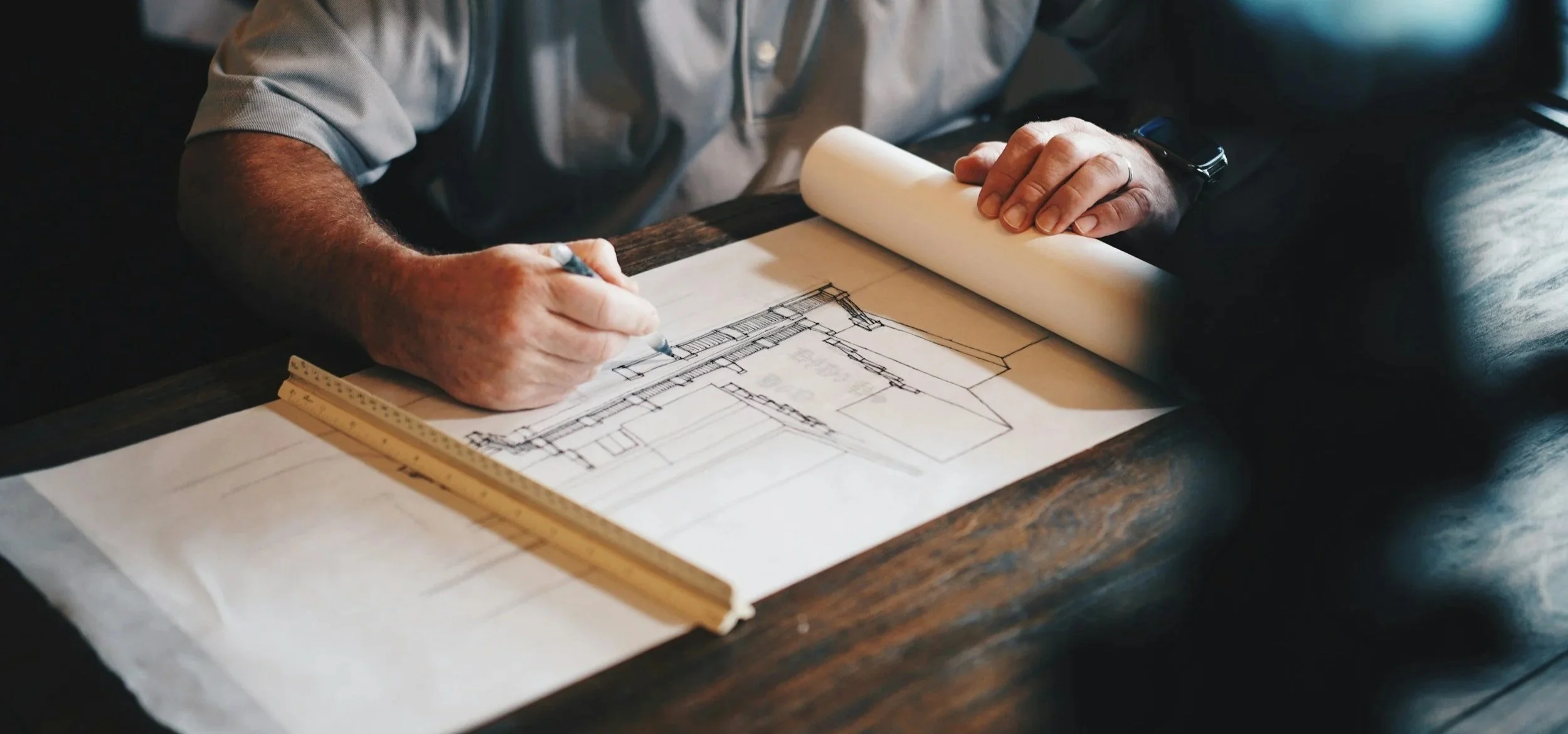 A person designing or drawing an architectural blueprint on paper, sitting at a dark wooden table.