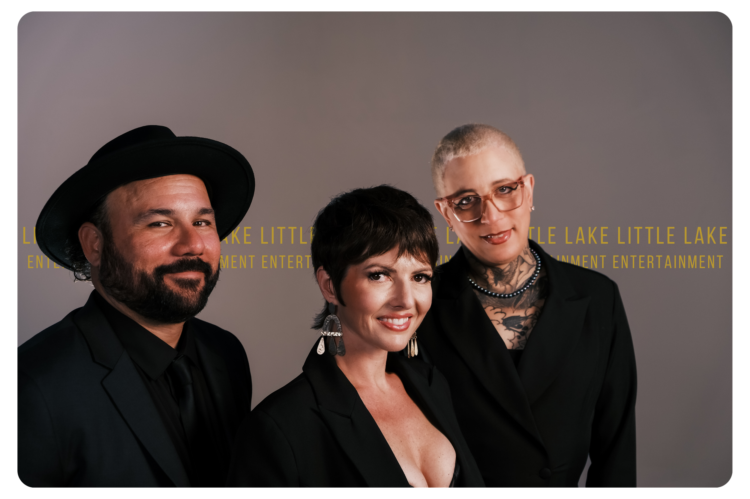 Three people posing for a photo, standing close together in front of a gray background with yellow text that reads 'LITTLE LAKE ENTERTAINMENT'.