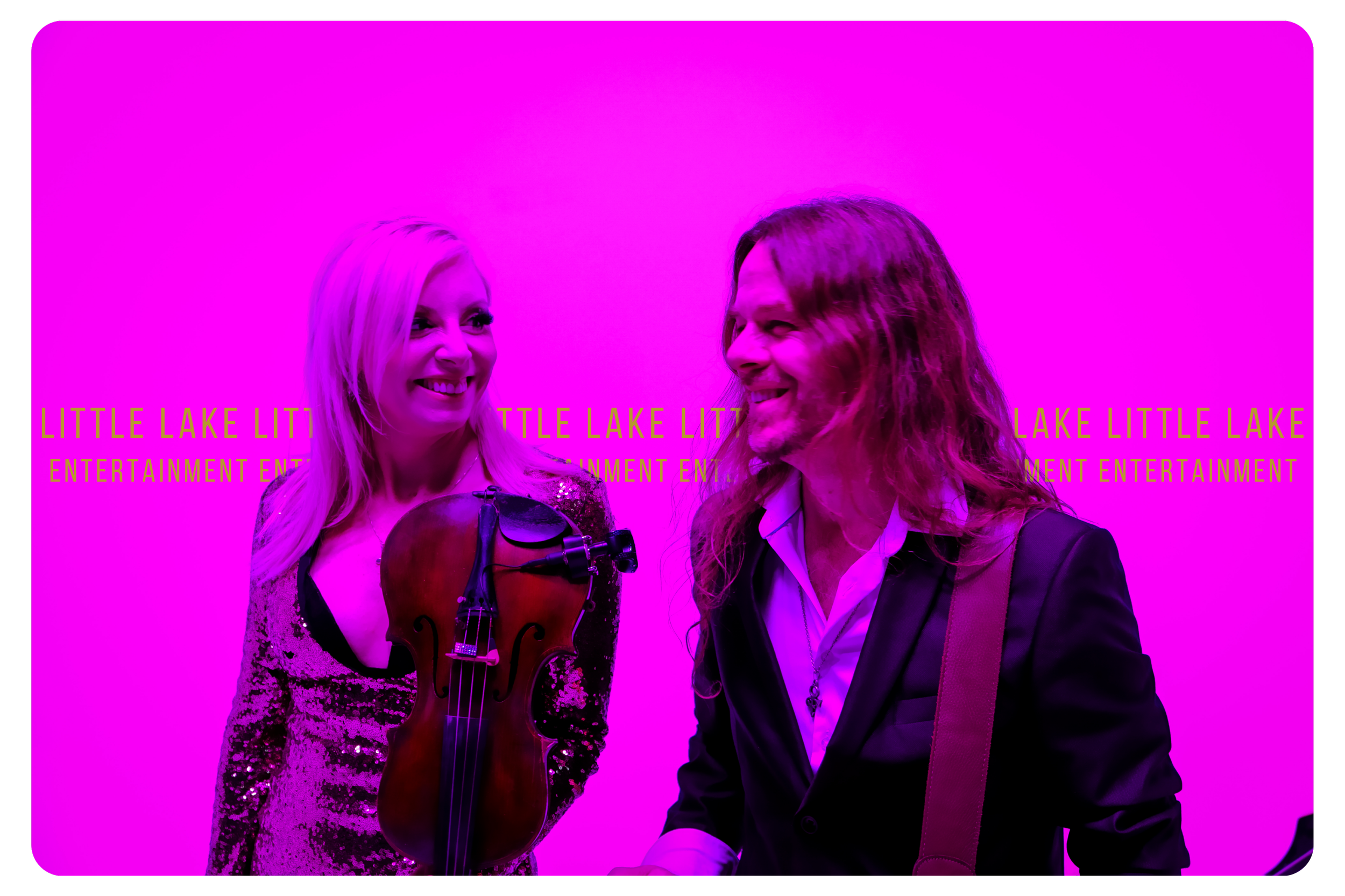 Two people smiling and talking at an event, with a bright pink background featuring the text 'Little Lake Little Lake Entertainment' repeatedly.