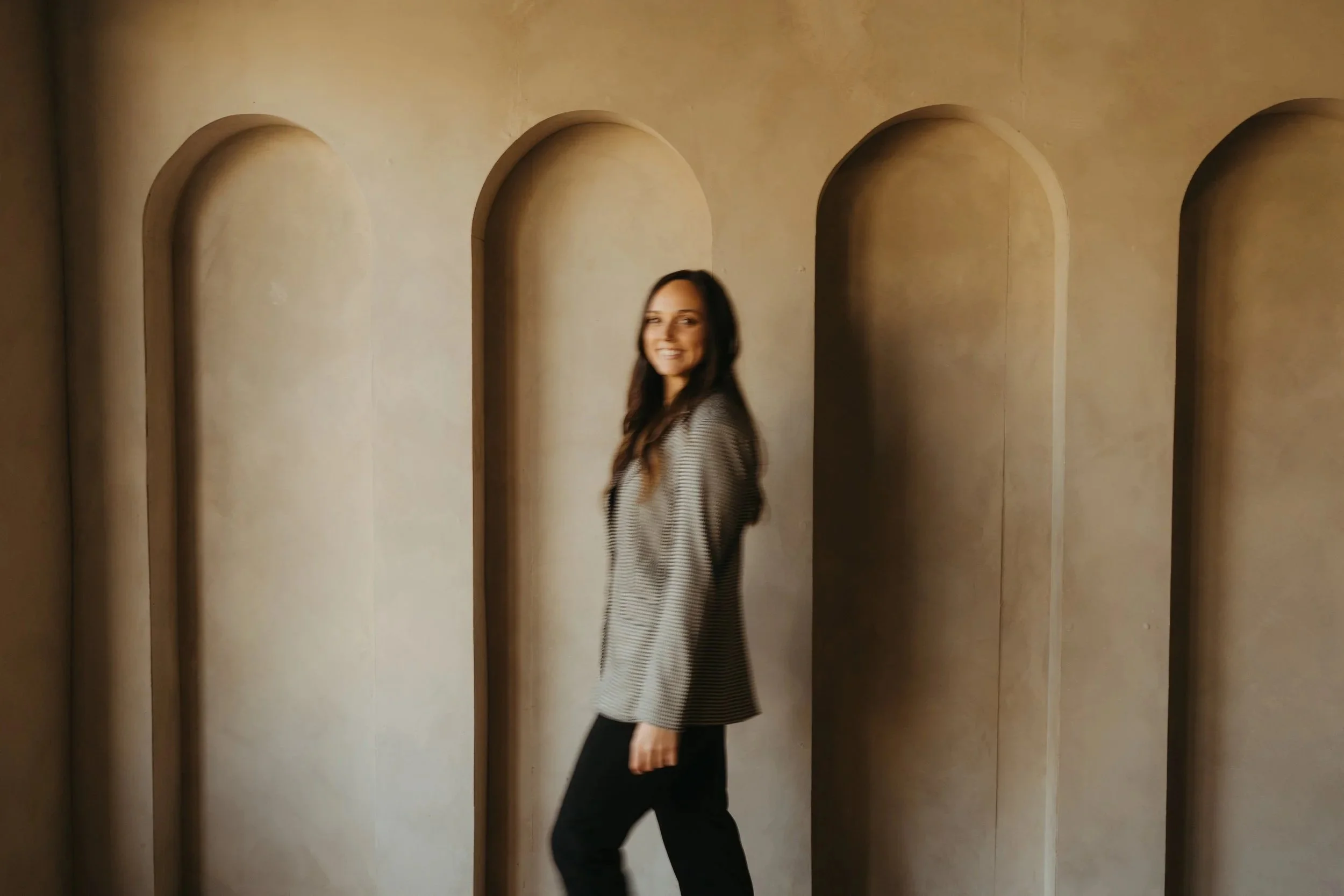 A woman with long brown hair smiling and walking in front of arched wall niches in beige color.