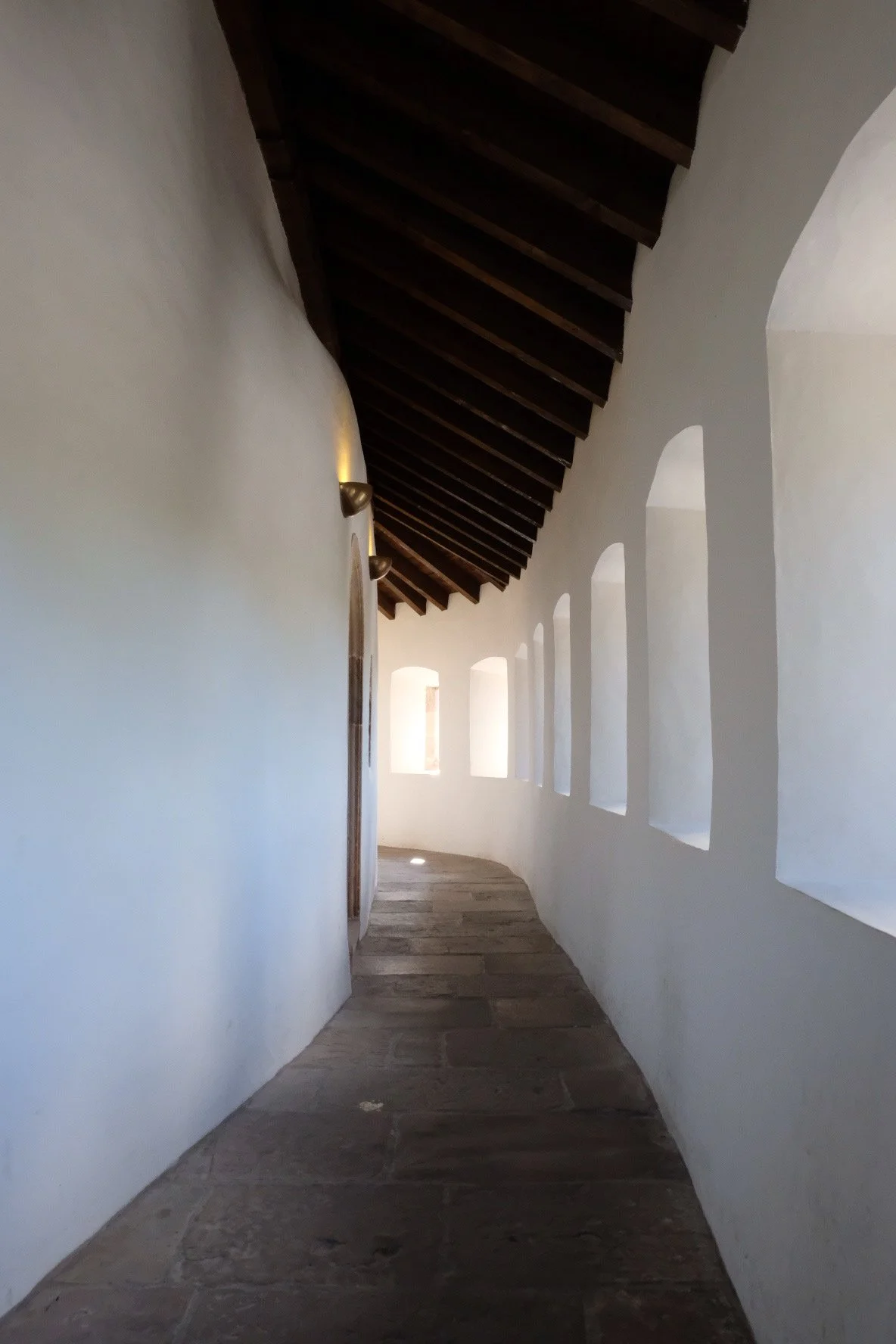 A narrow, curved hallway with white stucco walls, rustic stone flooring, and a dark wooden ceiling with exposed beams, illuminated by multiple small arched windows lining the right wall.