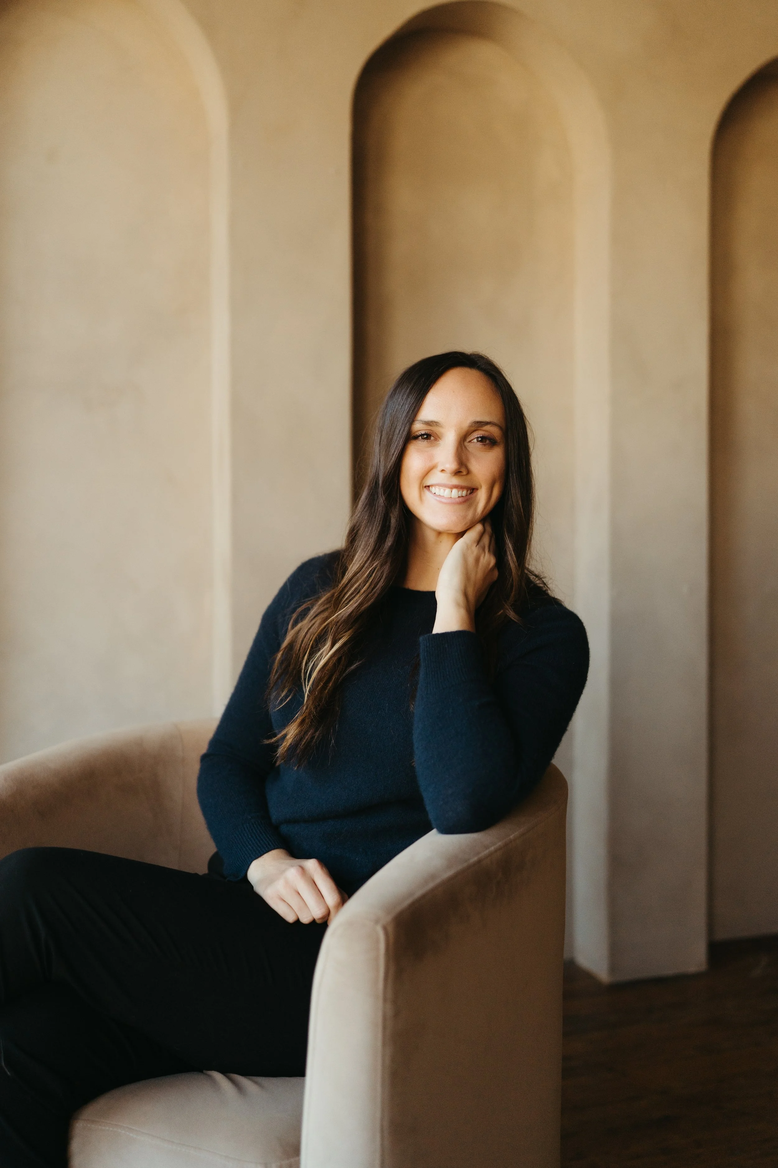 A woman with long dark hair wearing a black sweater sitting on a tan armchair, smiling with her hand near her neck, in front of a beige wall with arched alcoves.