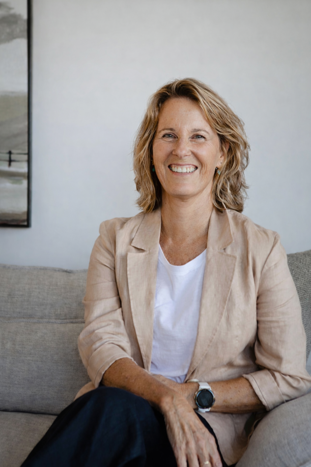 Smiling woman with shoulder-length curly hair, wearing a light beige blazer over a white shirt, sitting on a grey couch in a modern room.