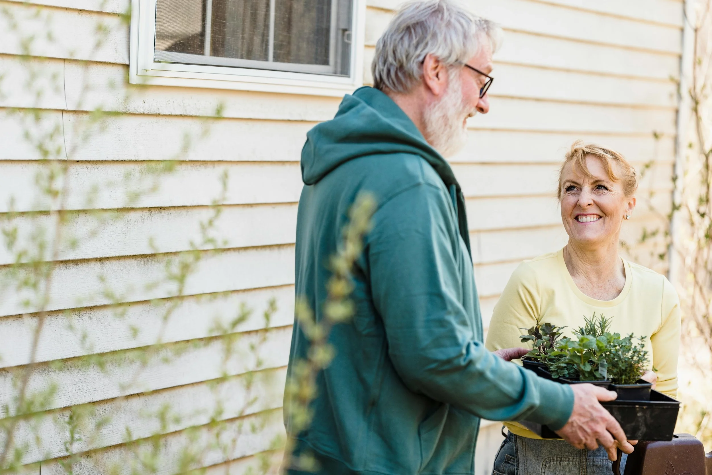 An elderly man and woman standing outside near a yellow wooden house, smiling and exchanging potted plants.