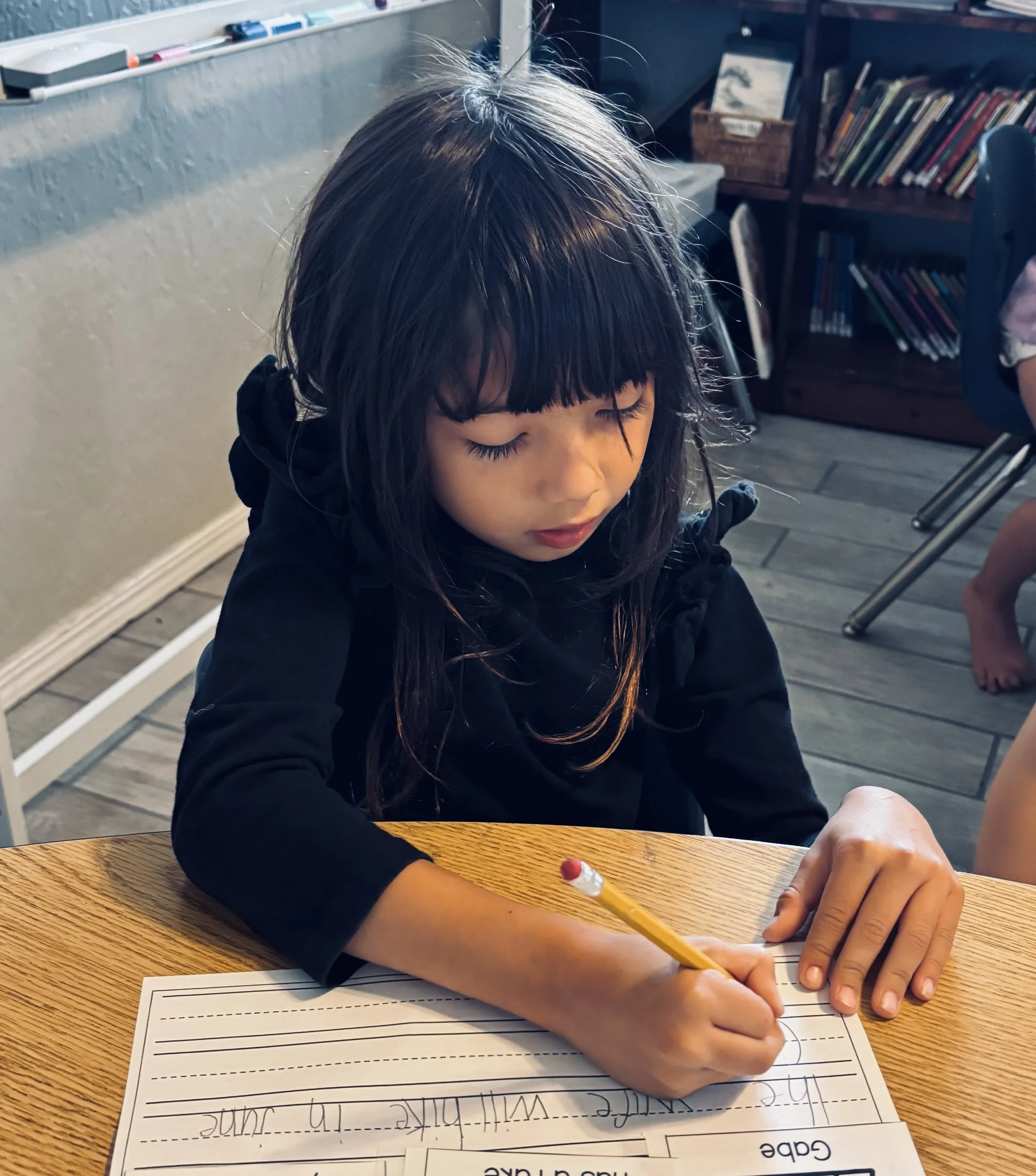 Young girl with black hair and bangs writing on a lined sheet of paper at a wooden table in a classroom.
