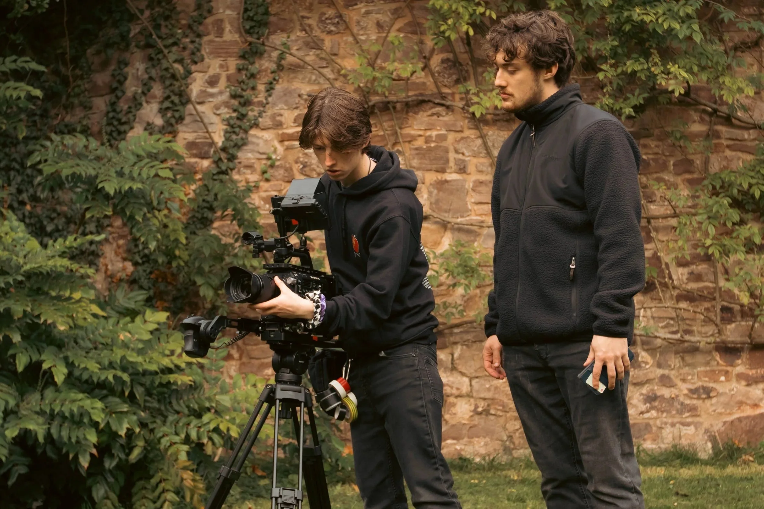 Two young men operating a professional camera on a tripod outdoors, with a stone wall and green foliage in the background.