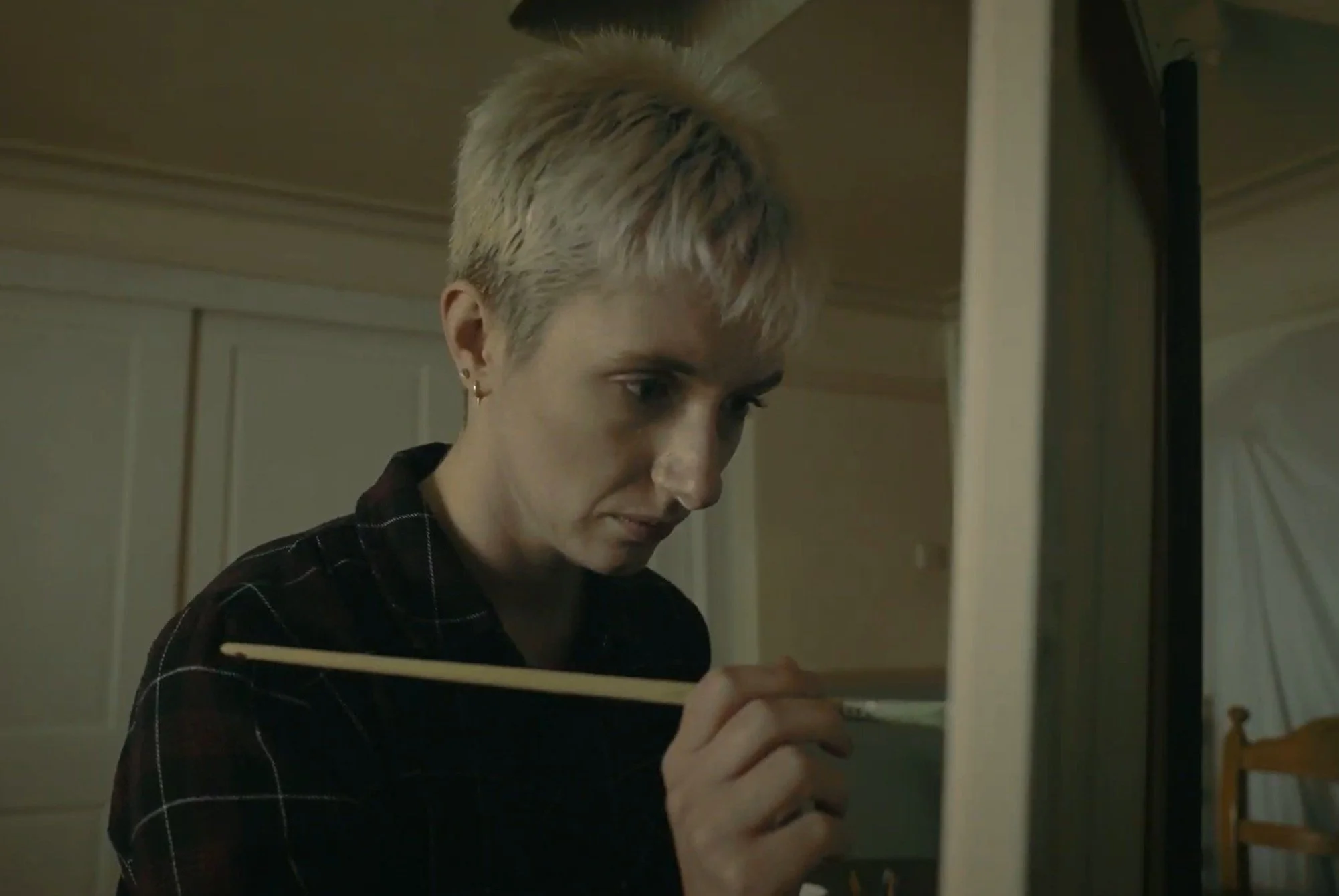 A young woman with short blond hair, wearing earrings and a dark plaid shirt, focused on painting on a canvas with a thin brush in a dimly lit room.