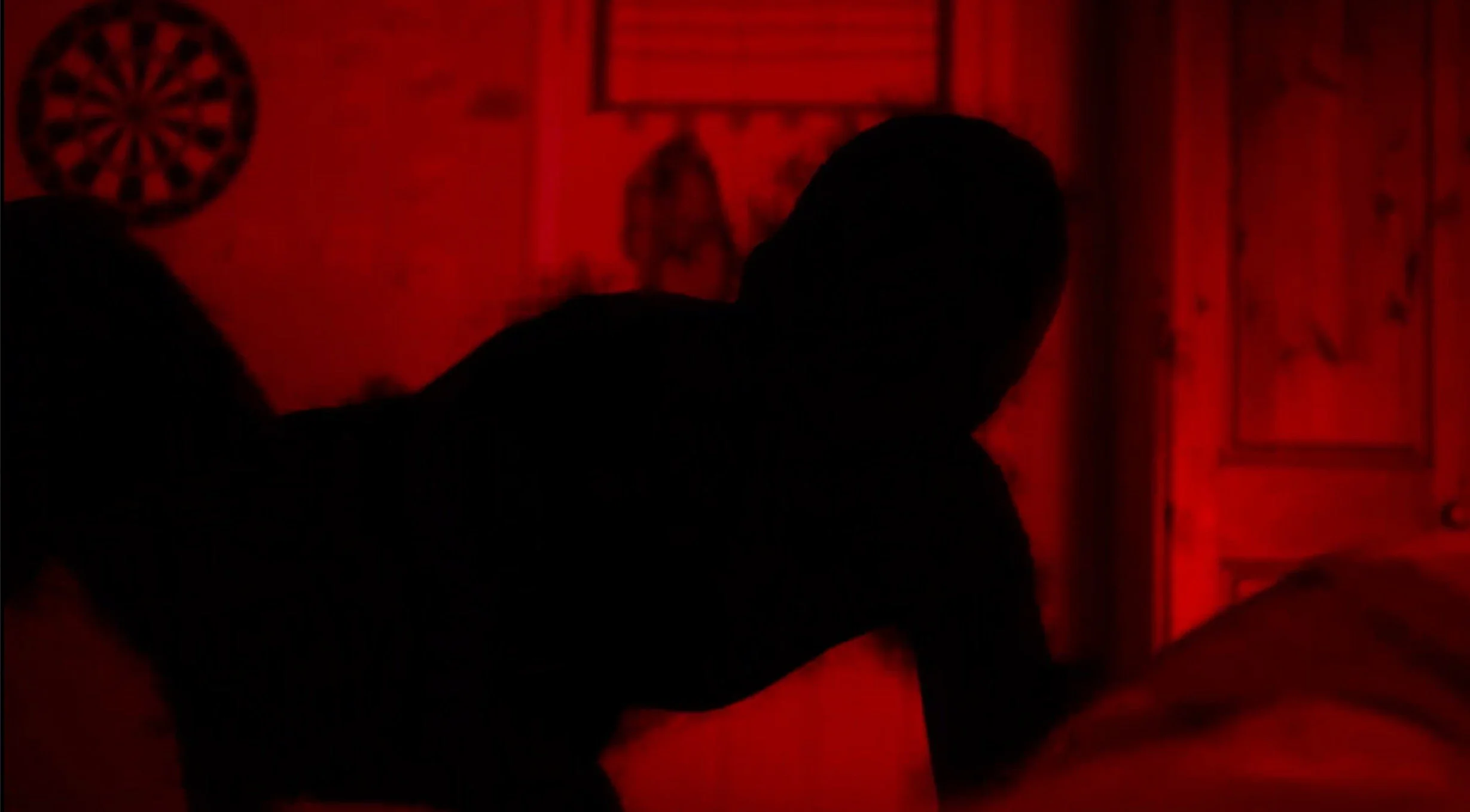 Silhouette of a person on a bed in a dimly lit room with red lighting, decorated with a dartboard on the wall.