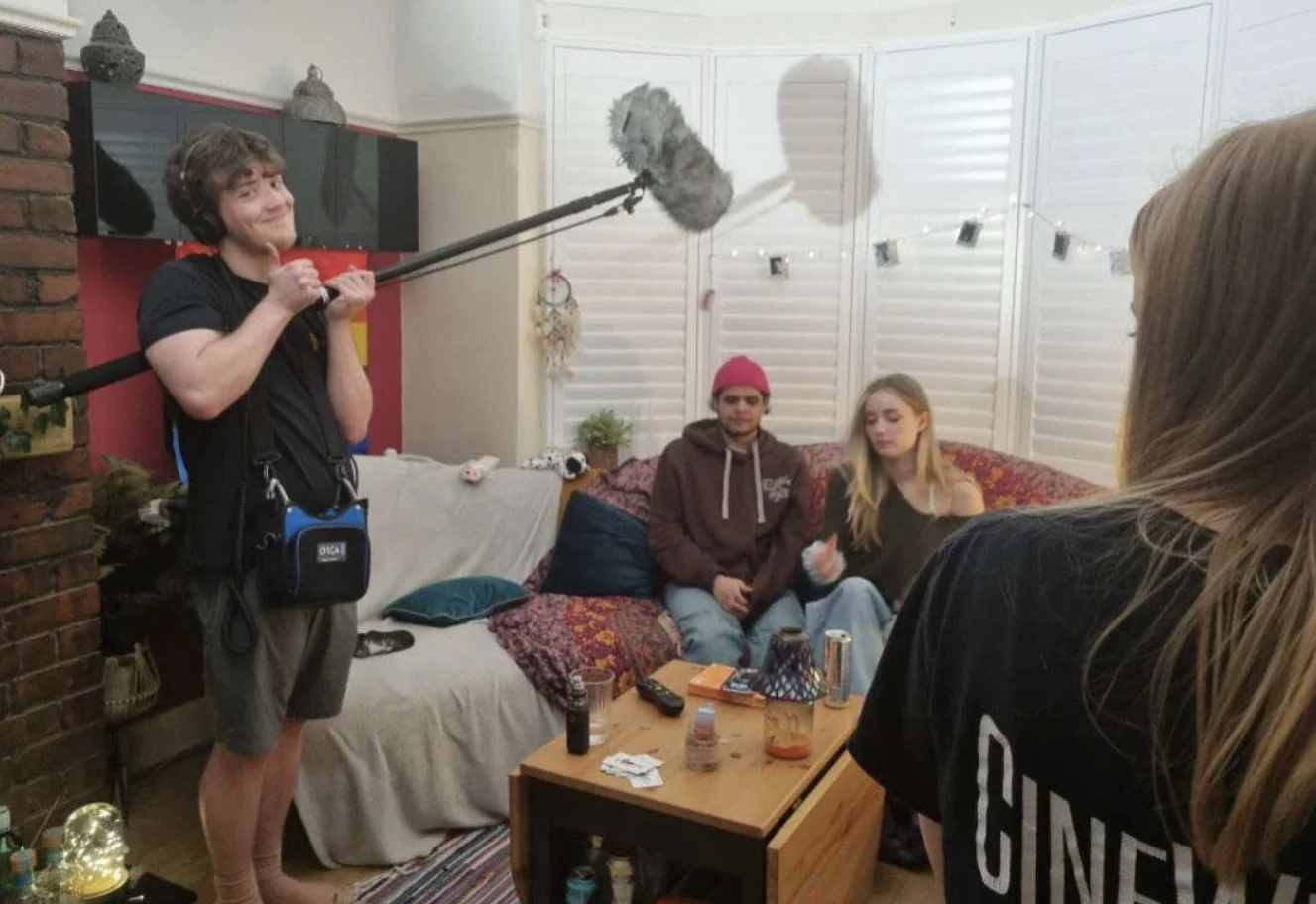 A person holding a boom microphone filming a group of three people sitting on a couch in a living room.