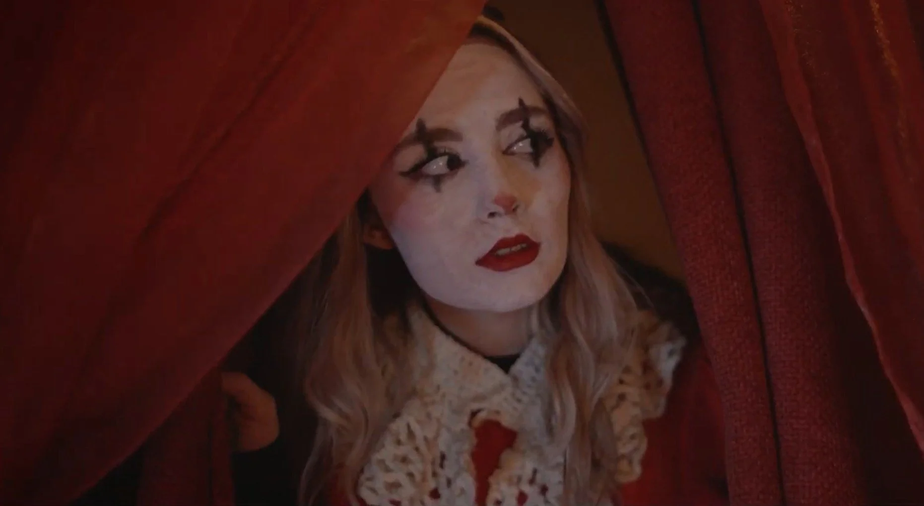Person peering out from behind red velvet curtains with dramatic makeup, including bold eyeliner and red lipstick.