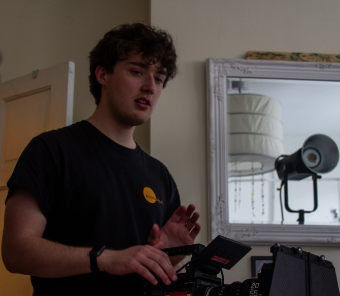 Young man with brown, curly hair operating a professional video camera indoors.
