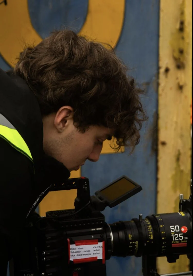 A person with curly brown hair looking through a professional camera lens, in an indoor setting with a yellow and blue background.