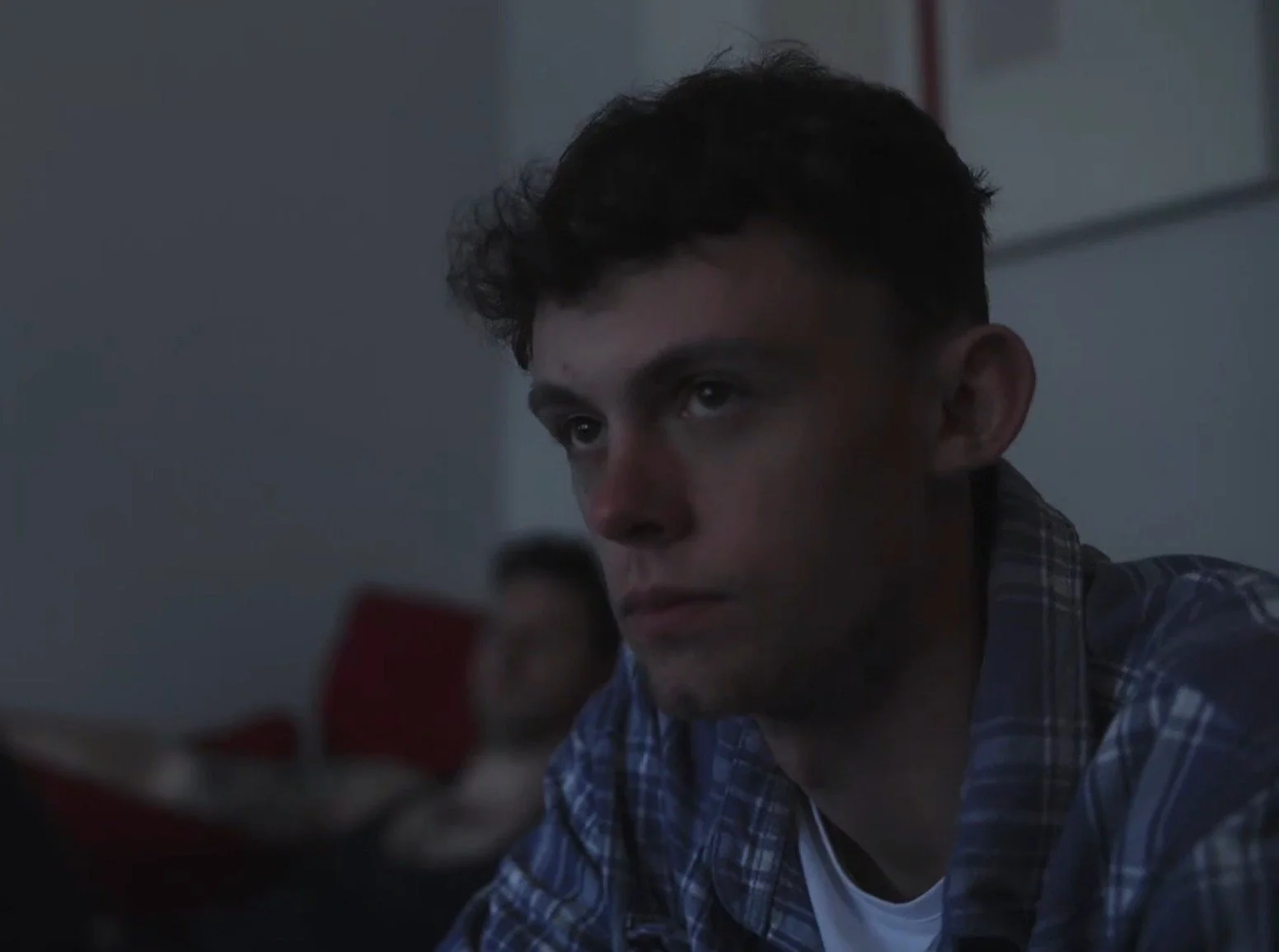 A young man with dark hair looking at his phone, with a woman blurred in the background, indoors in a dimly lit environment.
