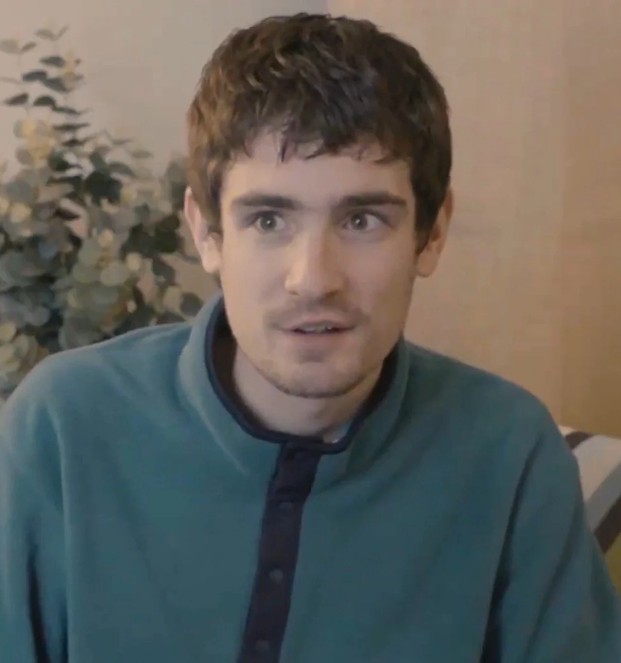 Young man with brown hair and blue eyes wearing a teal jacket, sitting indoors with a leafy plant in the background.