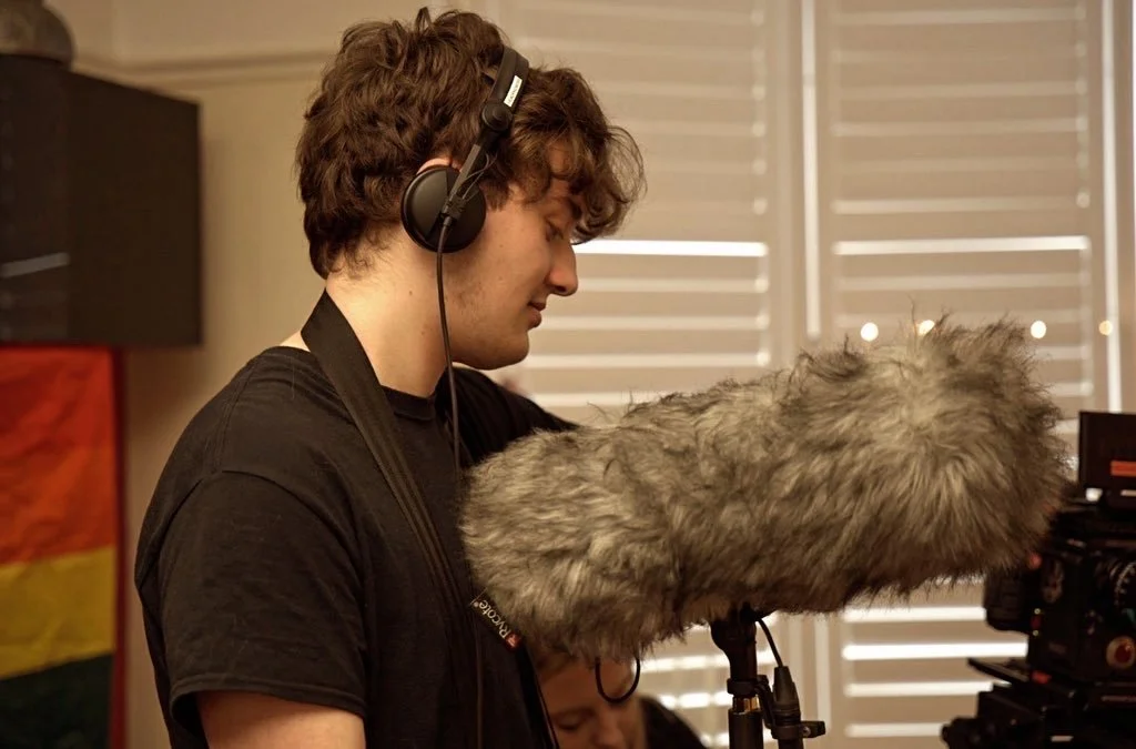 A young man wearing headphones standing behind a camera with a large furry windscreen, possibly filming or recording indoors, with a rainbow flag in the background.