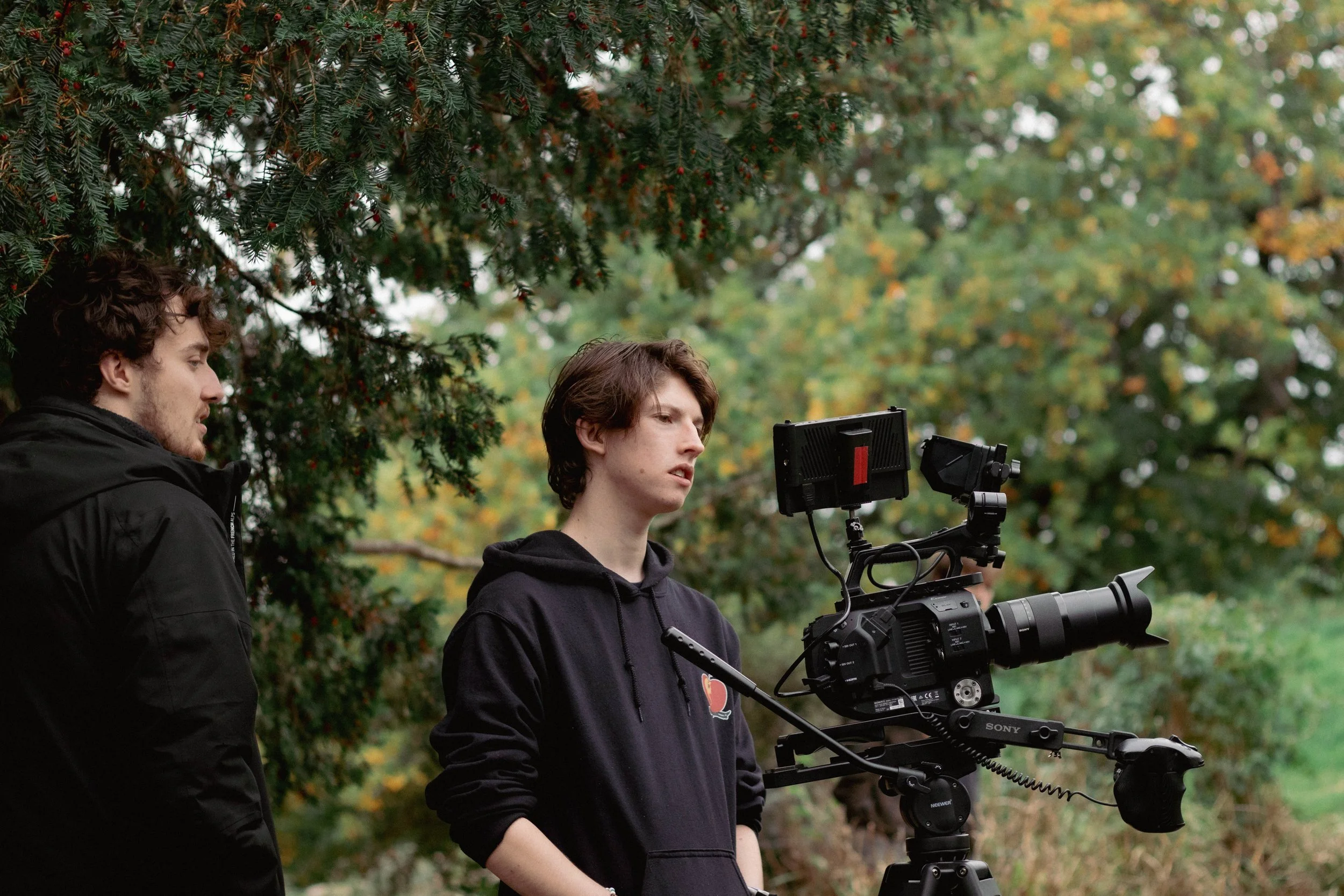 Two young men are outdoors in a wooded area, one is operating a professional video camera while the other observes.