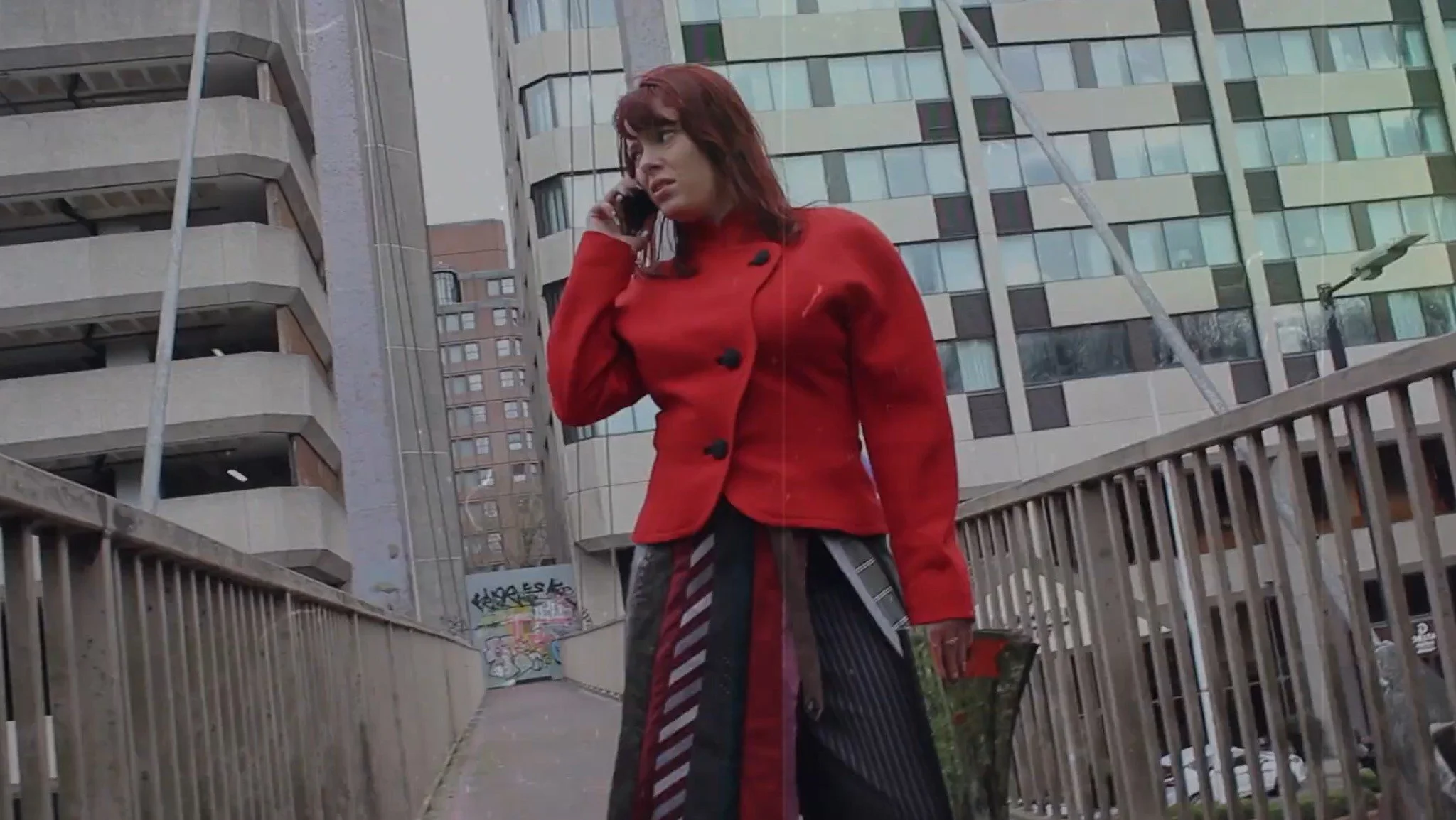 A person in a red jacket with a multicolored skirt standing on an urban bridge talking on a cellphone.