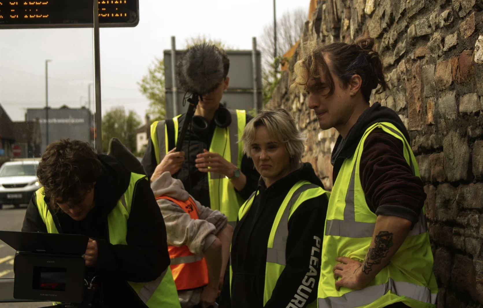 A group of people wearing yellow safety vests, gathered around a monitor outdoors, with a C-130 Hercules aircraft in the blurred background and a stone wall on one side.