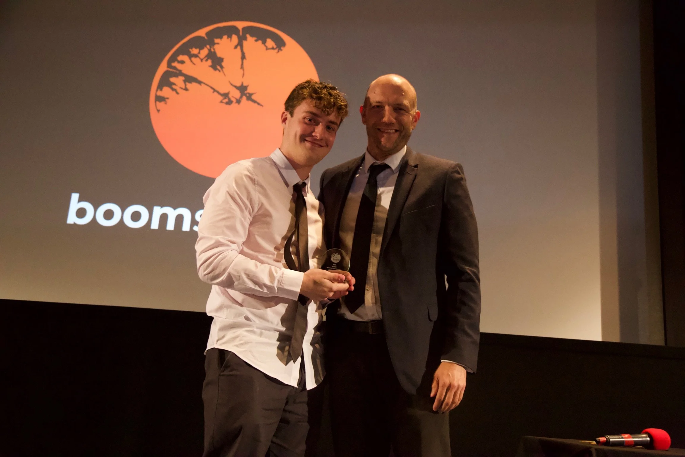 Two men in suits standing on stage after an award ceremony, smiling, with a booms logo projected behind them.