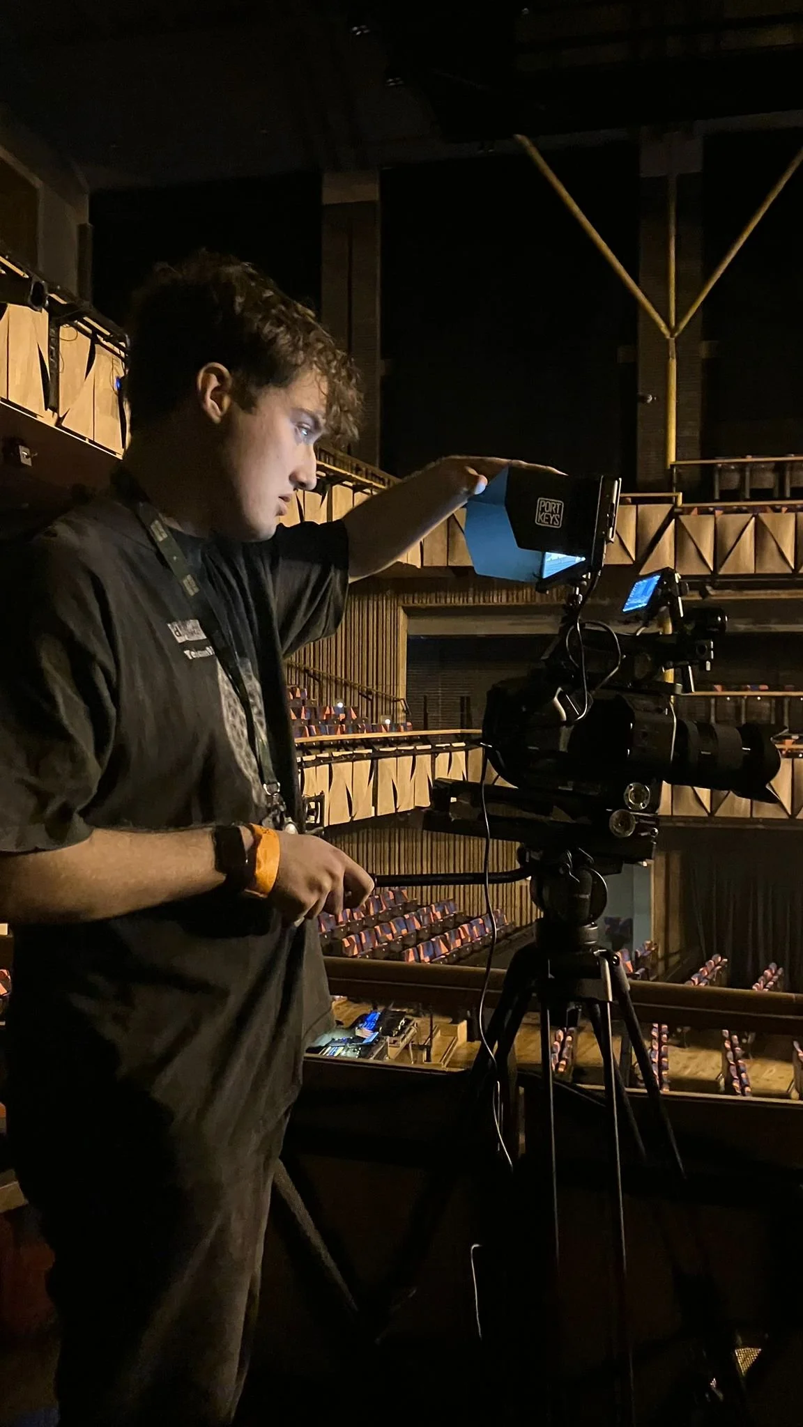A young man adjusting a video camera on a tripod in an empty theater or auditorium.