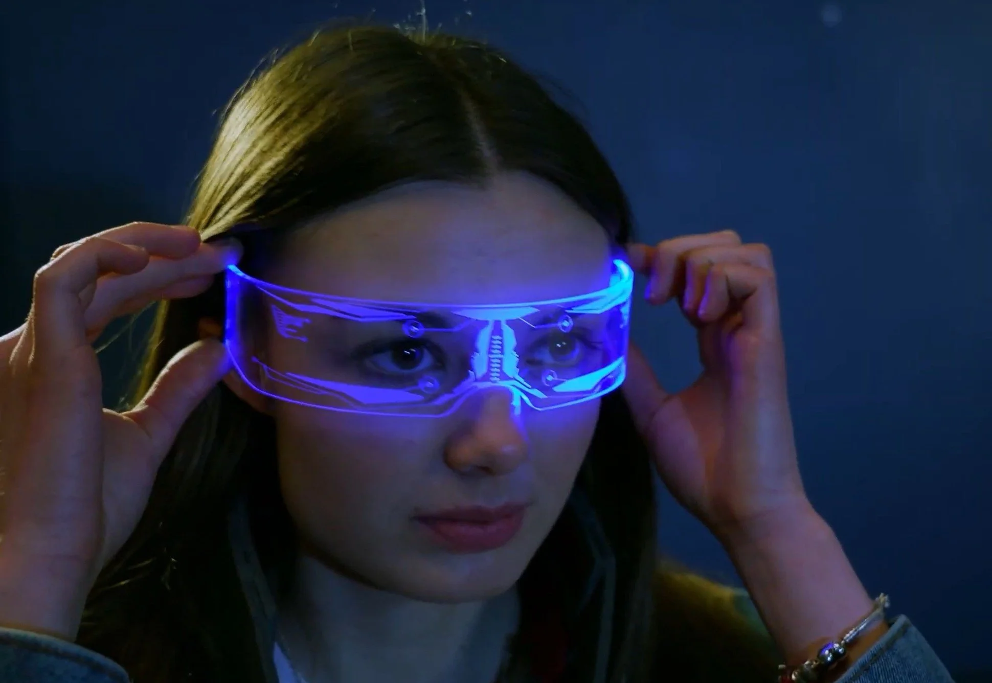 A woman with long brown hair wearing futuristic blue LED glasses with a dark background.
