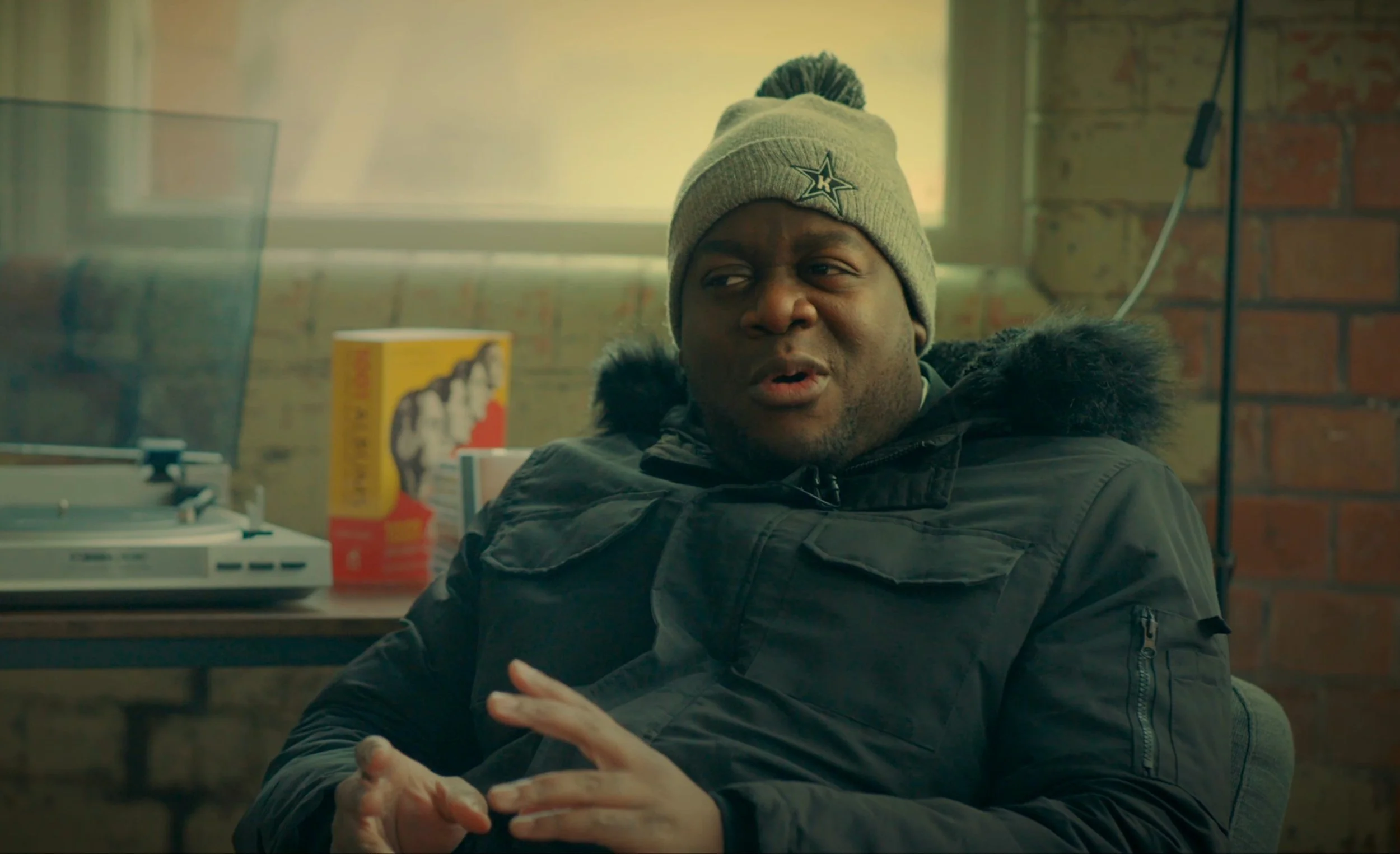 A man wearing a gray beanie with a star logo and a black winter coat sitting in a room with a brick wall. There are books and a turntable on a table behind him.