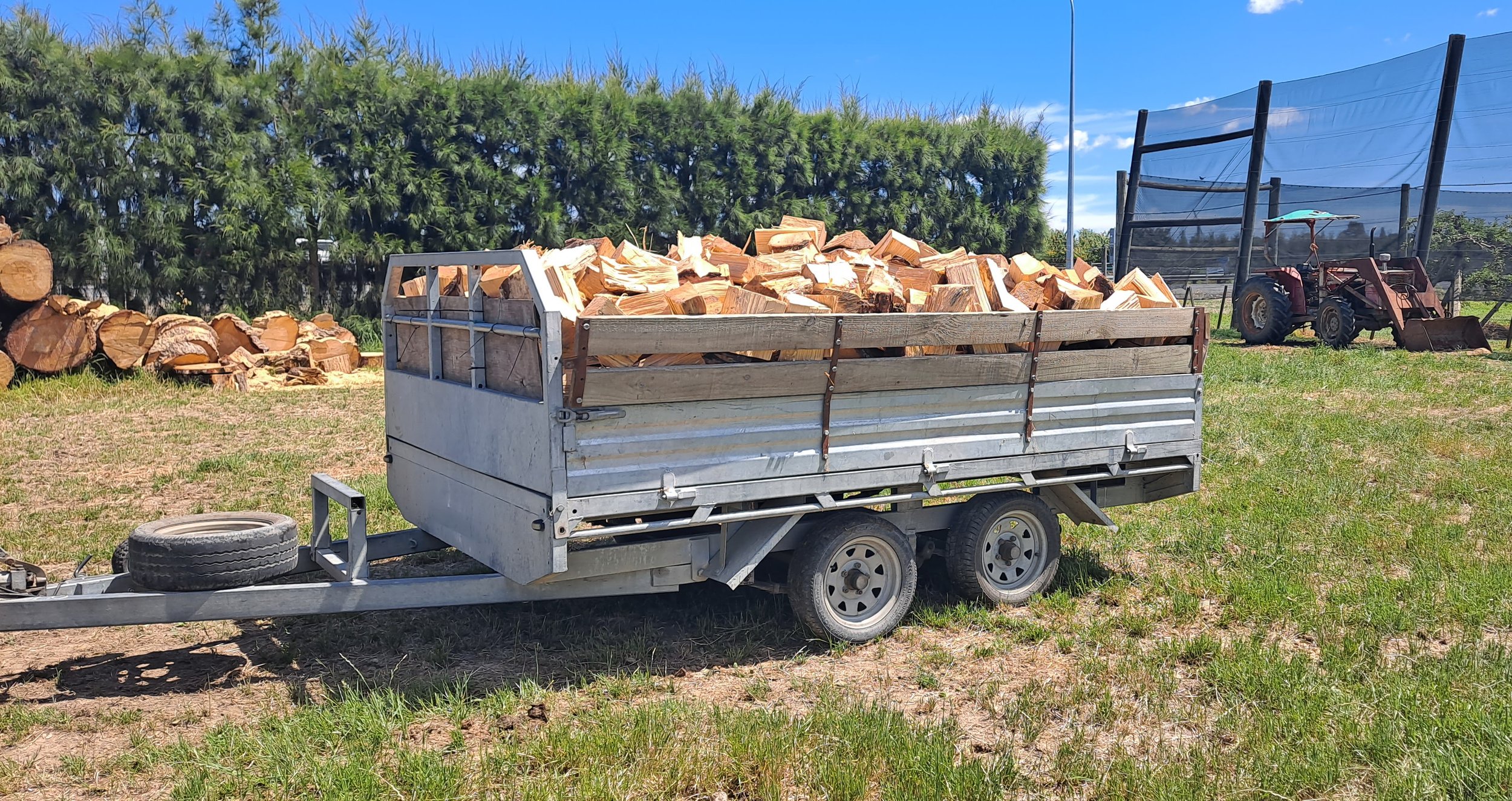 Trailer filled with chopped firewood parked on grass with additional logs and a tractor in the background.