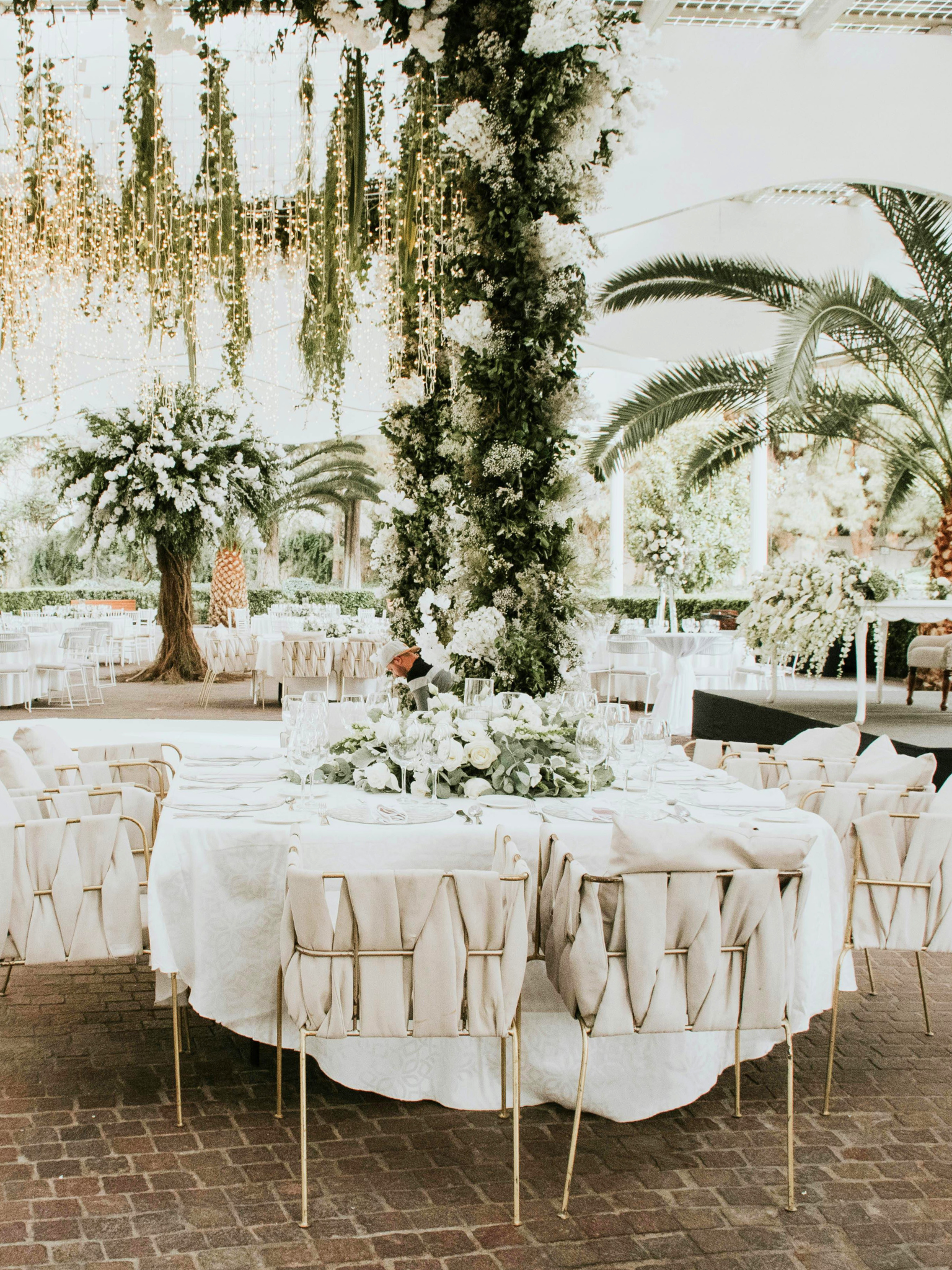 Mesas decoradas con flores blancas y vajilla para un evento, en un espacio decorado con plantas verdes y flores, probablemente una boda o evento formal.
