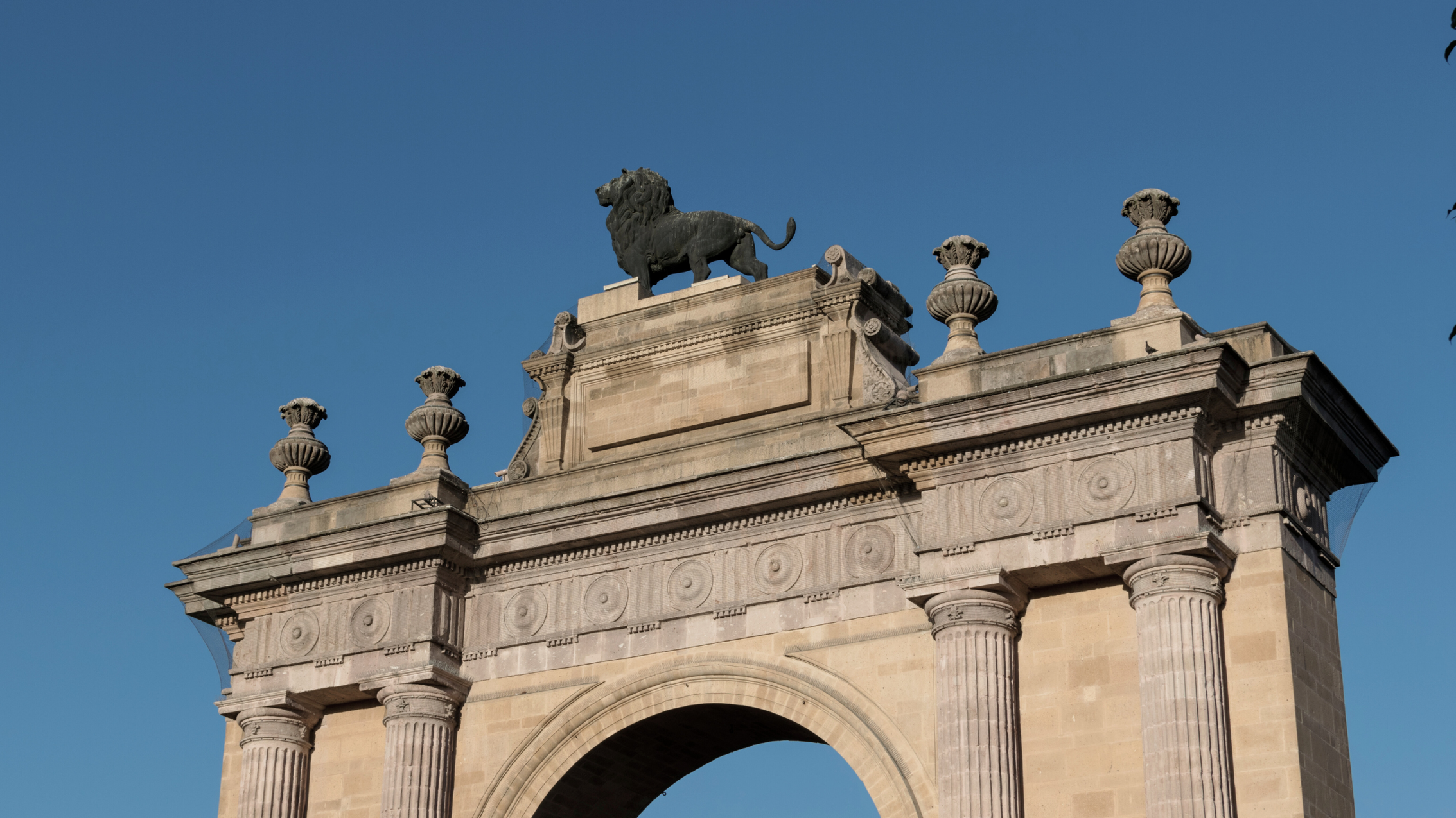 Detalle de la parte superior de un monumento con una escultura de un león en la cima, contra un cielo azul claro.