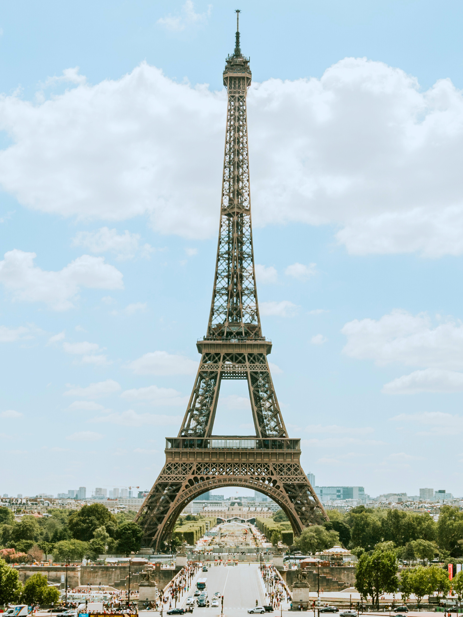 La Torre Eiffel vista desde los jardines de Champ de Mars, con muchas personas y coches en primer plano y el cielo con nubes en segundo plano.