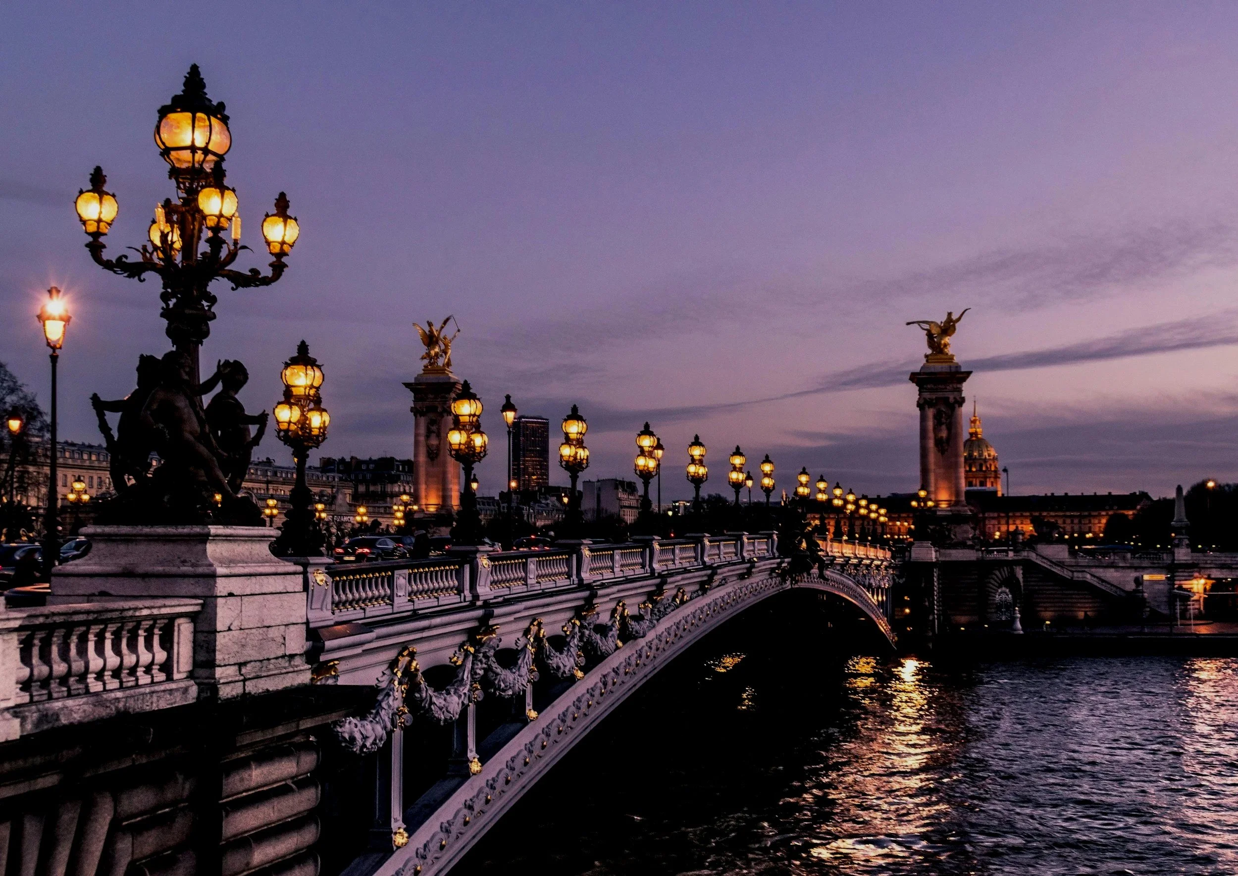 Puente con farolas encendidas al atardecer, con estatuas de ángeles en las pilas y un edificio emblemático con cúpula dorada al fondo.