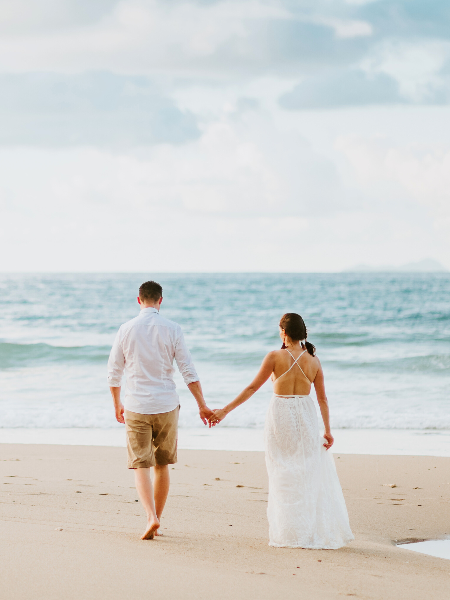 Pareja caminando por la orilla del mar con el hombre vestido con camisa blanca y pantalones cortos beige, y la mujer con un vestido largo y blanco. Ambos sosteniéndose de las manos mientras caminan hacia el agua.