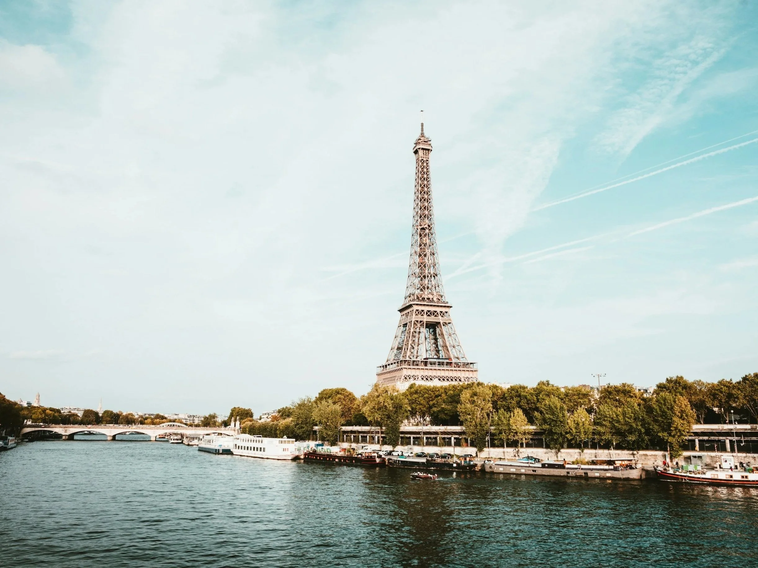 La Torre Eiffel vista desde el río Sena en París, con barcos y árboles en primer plano y un cielo despejado.