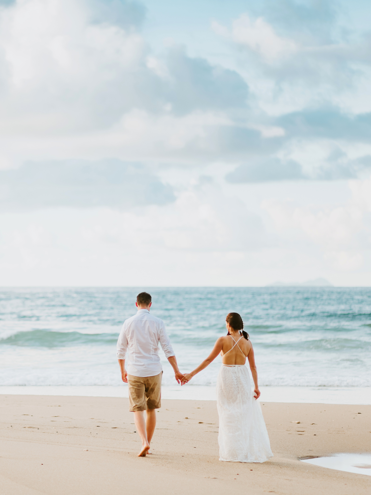 Una pareja caminando por la playa de la mano, vista desde atrás, con el mar y el cielo con nubes de fondo.