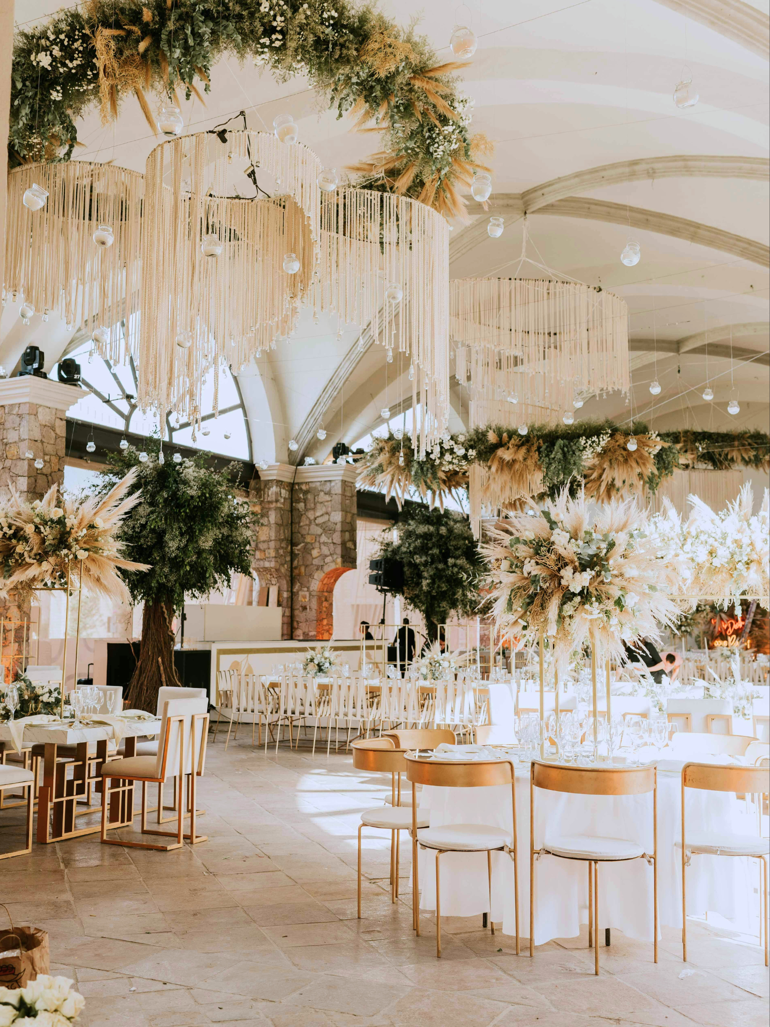 Decoración de evento con arreglos florales, centros de mesa, cortinas de mimbre y lámparas colgantes, en un espacio amplio con techo arqueado.