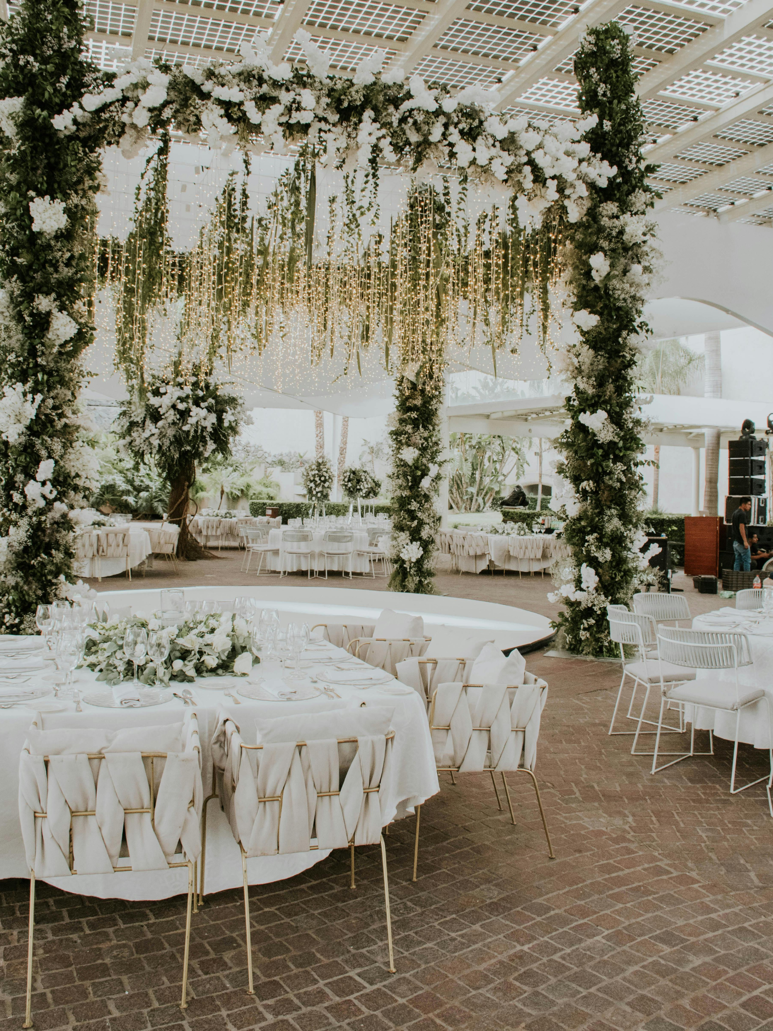 Arco decorativo de flores blancas y verdes con luces colgantes, en un espacio de evento elegante con mesas y sillas blancas, techo de madera y paredes blancas.