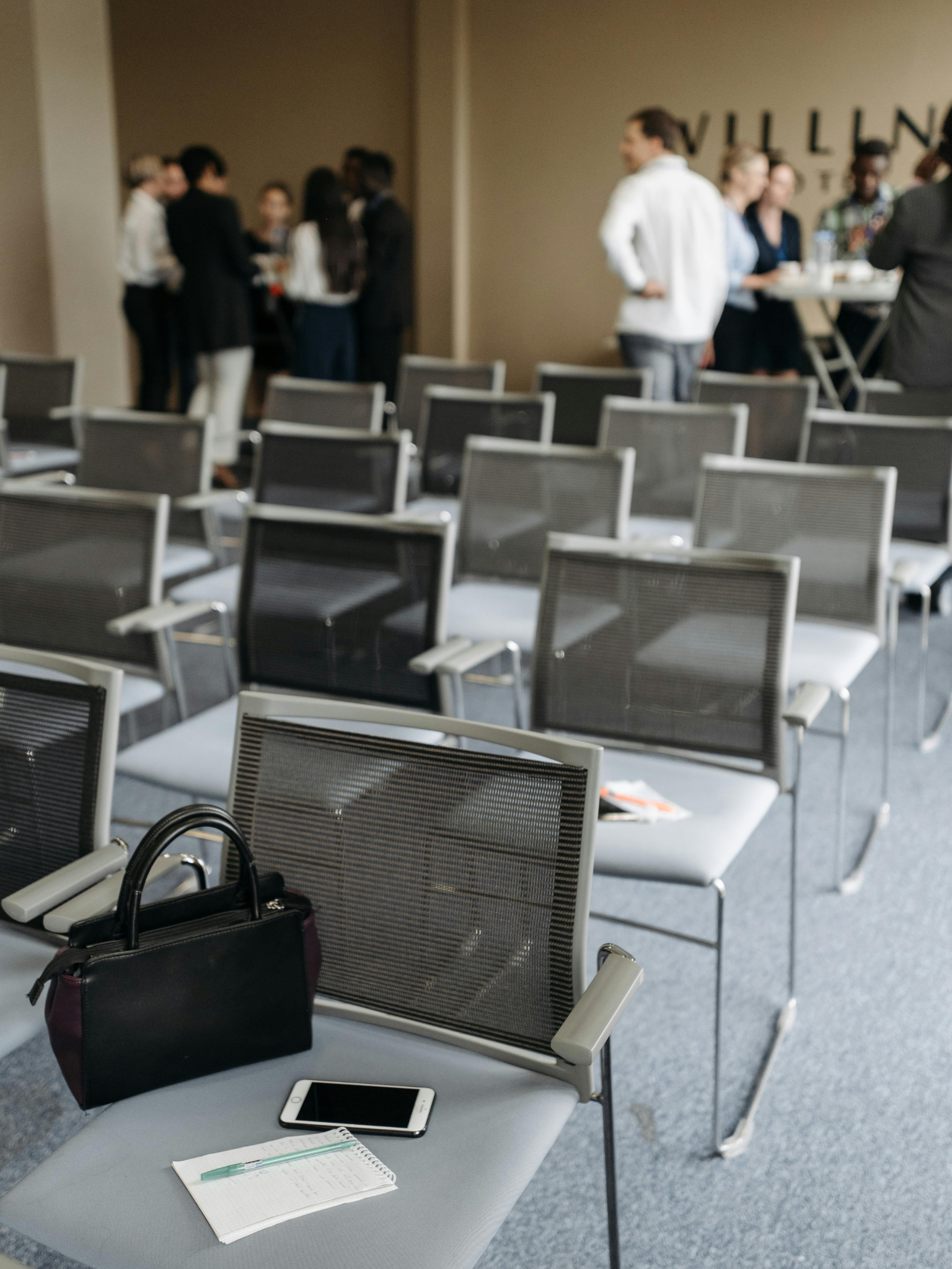 Sillas vacías en un auditorio, con una mesa en primer plano que tiene una libreta, bolígrafo, teléfono móvil, bolso y otros objetos, y en el fondo varias personas conversando en un pasillo