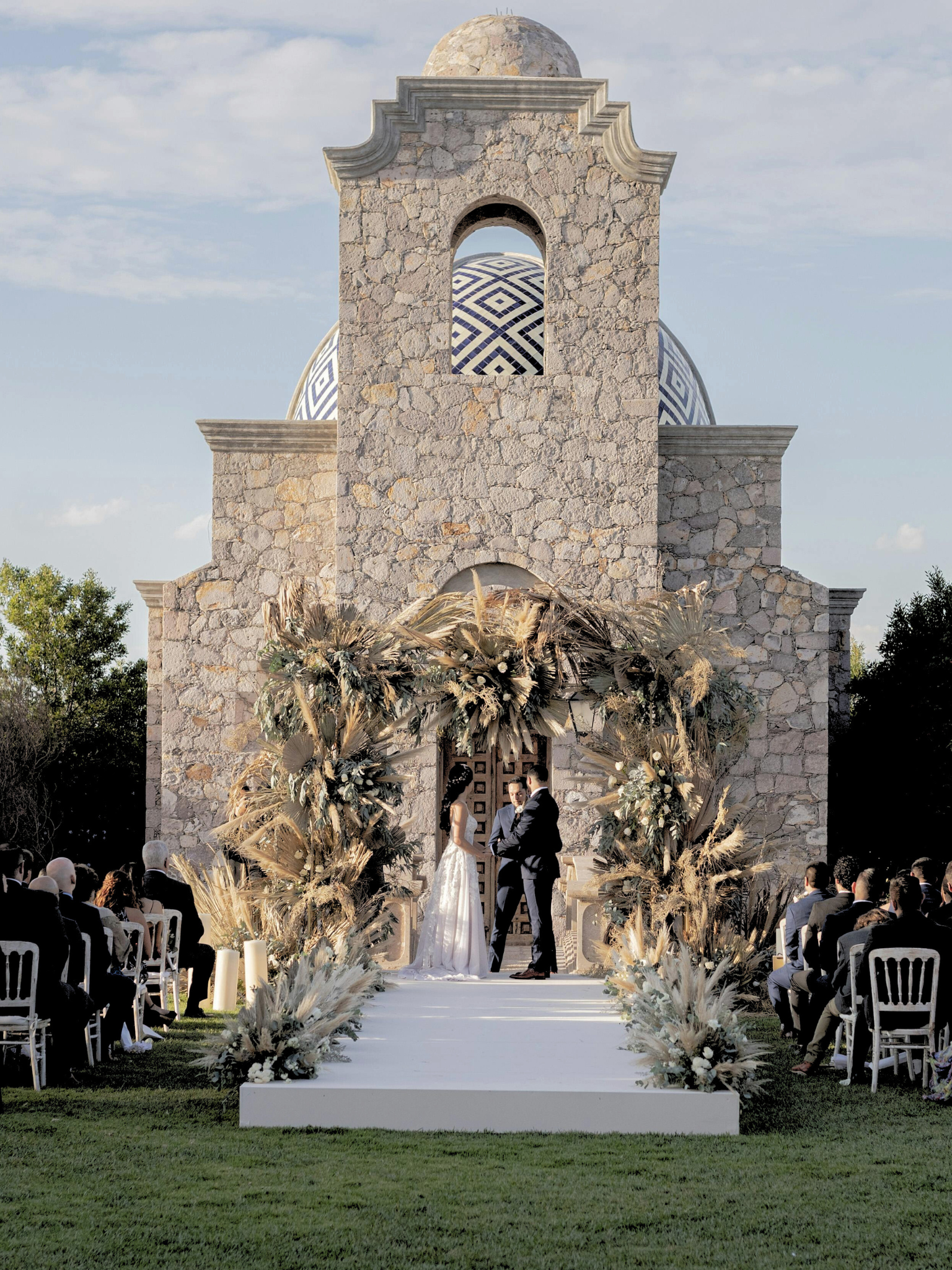 Boda en una iglesia con decoraciones con follaje y flores en tonos neutros, con una pareja intercambiando votos frente a un altar y asistentes sentados en ambos lados de la pasarela.